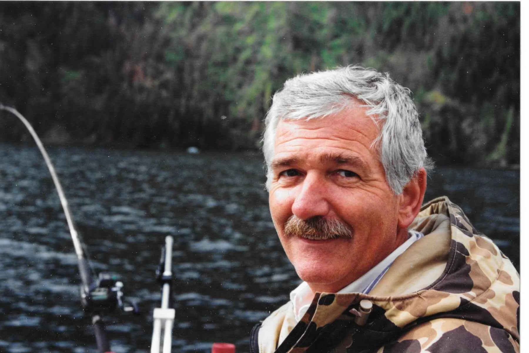 Man with gray hair wearing a camo jacket near a body of water, smiling at the camera, with fishing gear in the background.