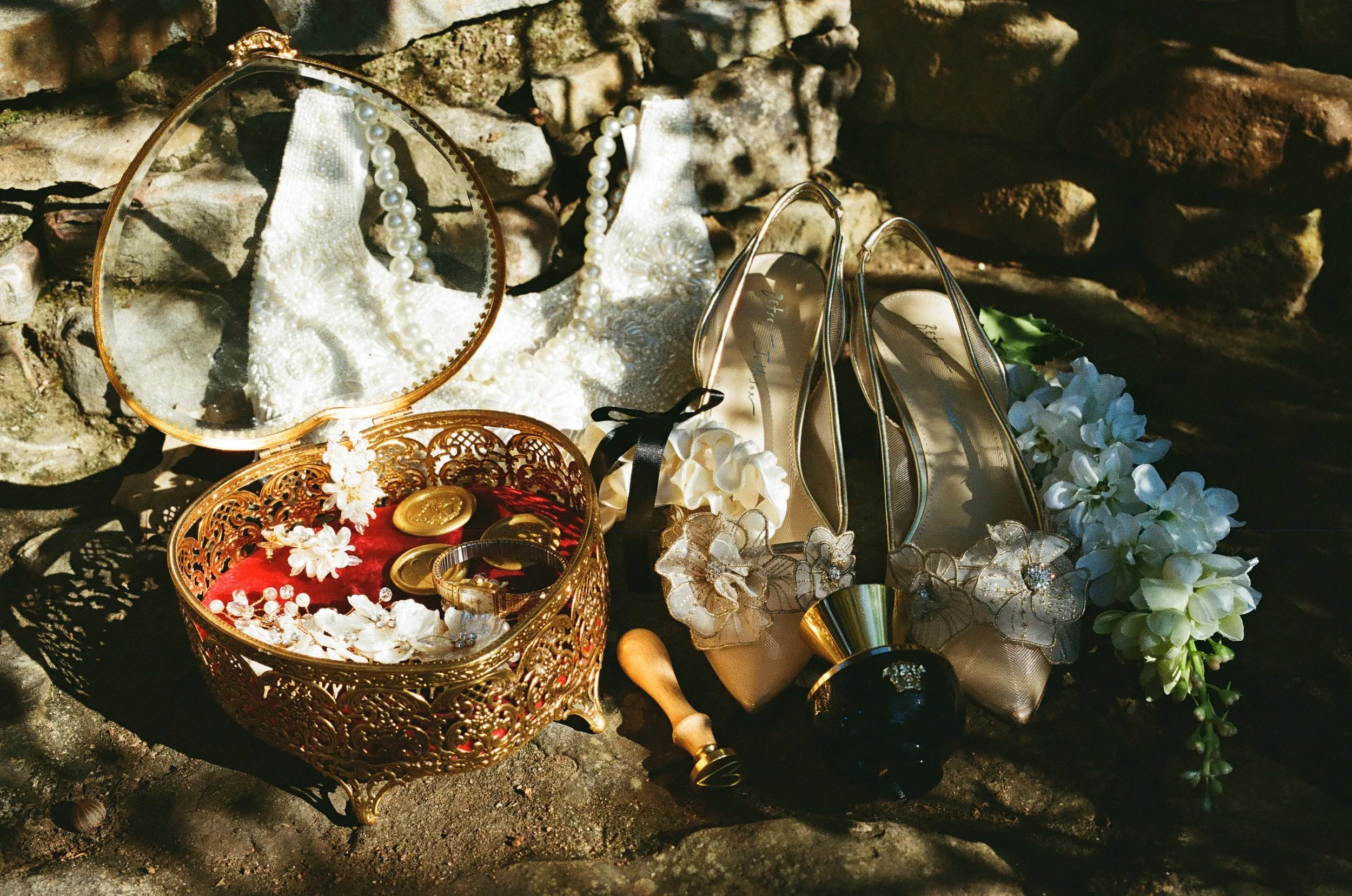 A collection of elegant wedding accessories including a pearl necklace, a pair of gold high heels with floral embellishments, a white ribbon, a small mirror, and a decorative box with jewelry, all arranged on a stone surface against a stone wall back