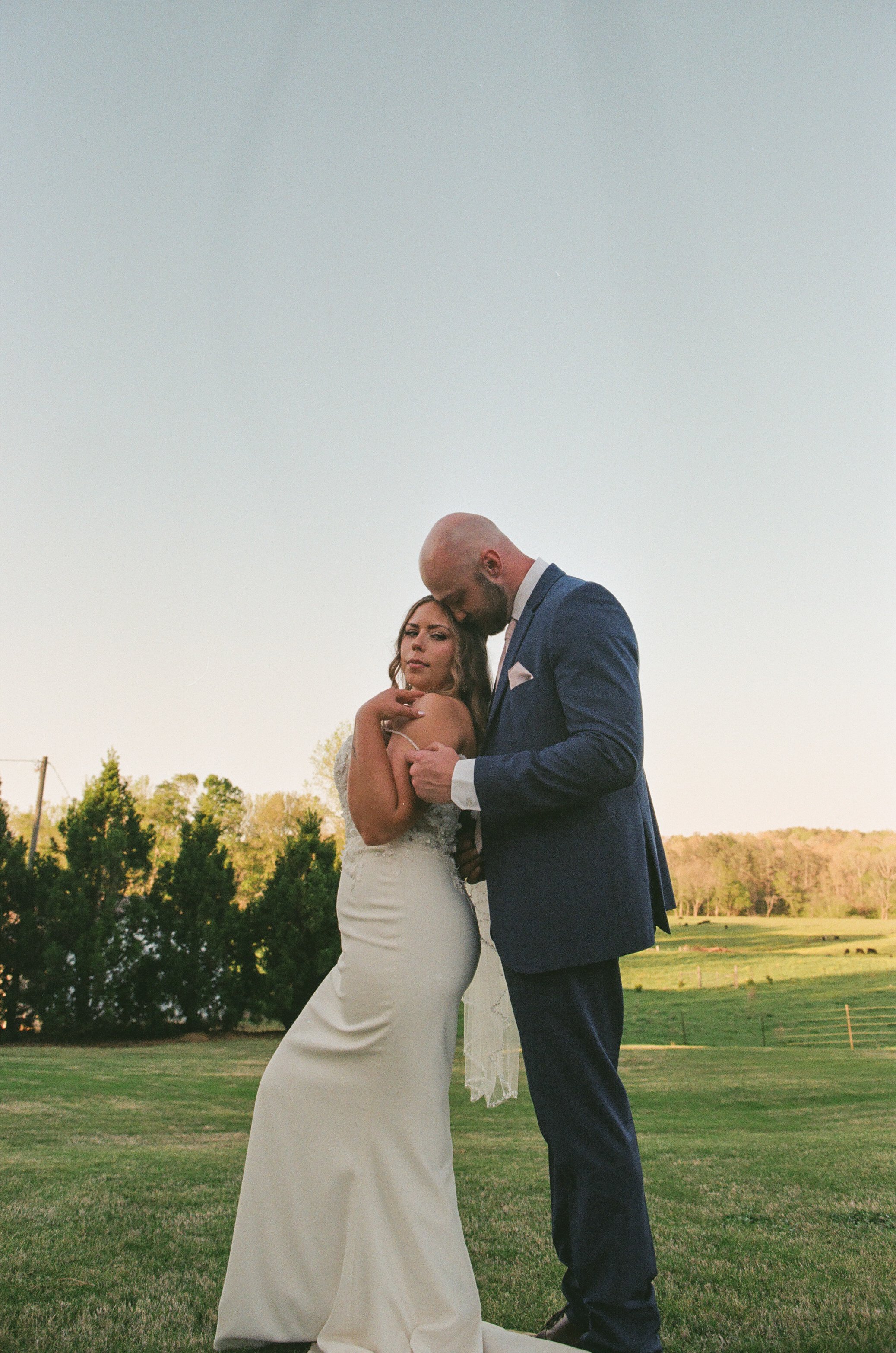 A bride and groom standing close together on a grassy field during sunset, with trees in the background. The bride is in a white wedding dress, and the groom is in a navy suit. The groom has his head bowed toward the bride, who looks at the camera at