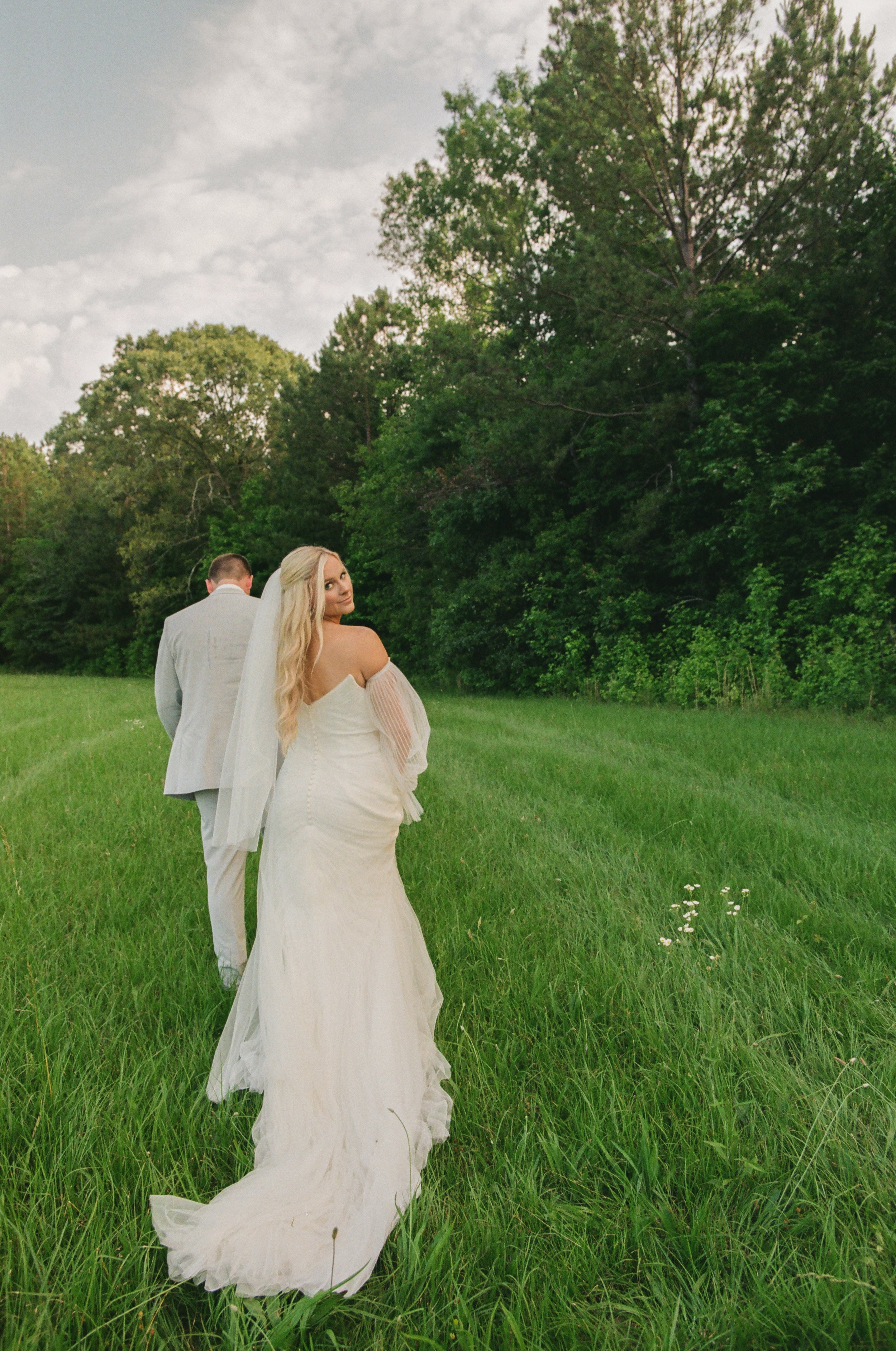 A bride in a wedding dress and a veil walking in a grassy field, with a groom in a gray suit ahead of her, surrounded by trees and a partly cloudy sky.