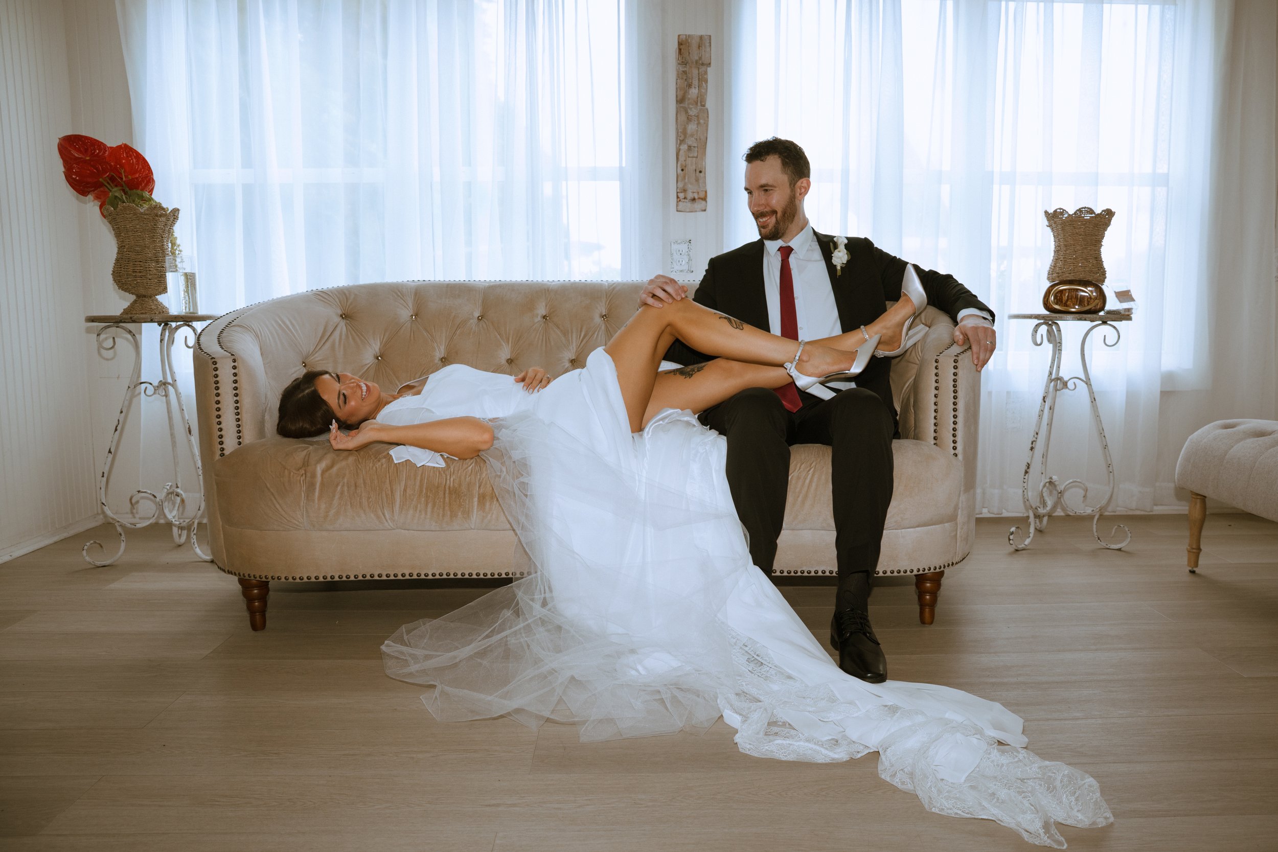 A bride lying on a beige tufted sofa with her legs raised and crossed, wearing a white wedding dress, and a groom sitting beside her, smiling and holding her leg, dressed in a black suit with a red tie, in a bright, airy room with sheer white curtain