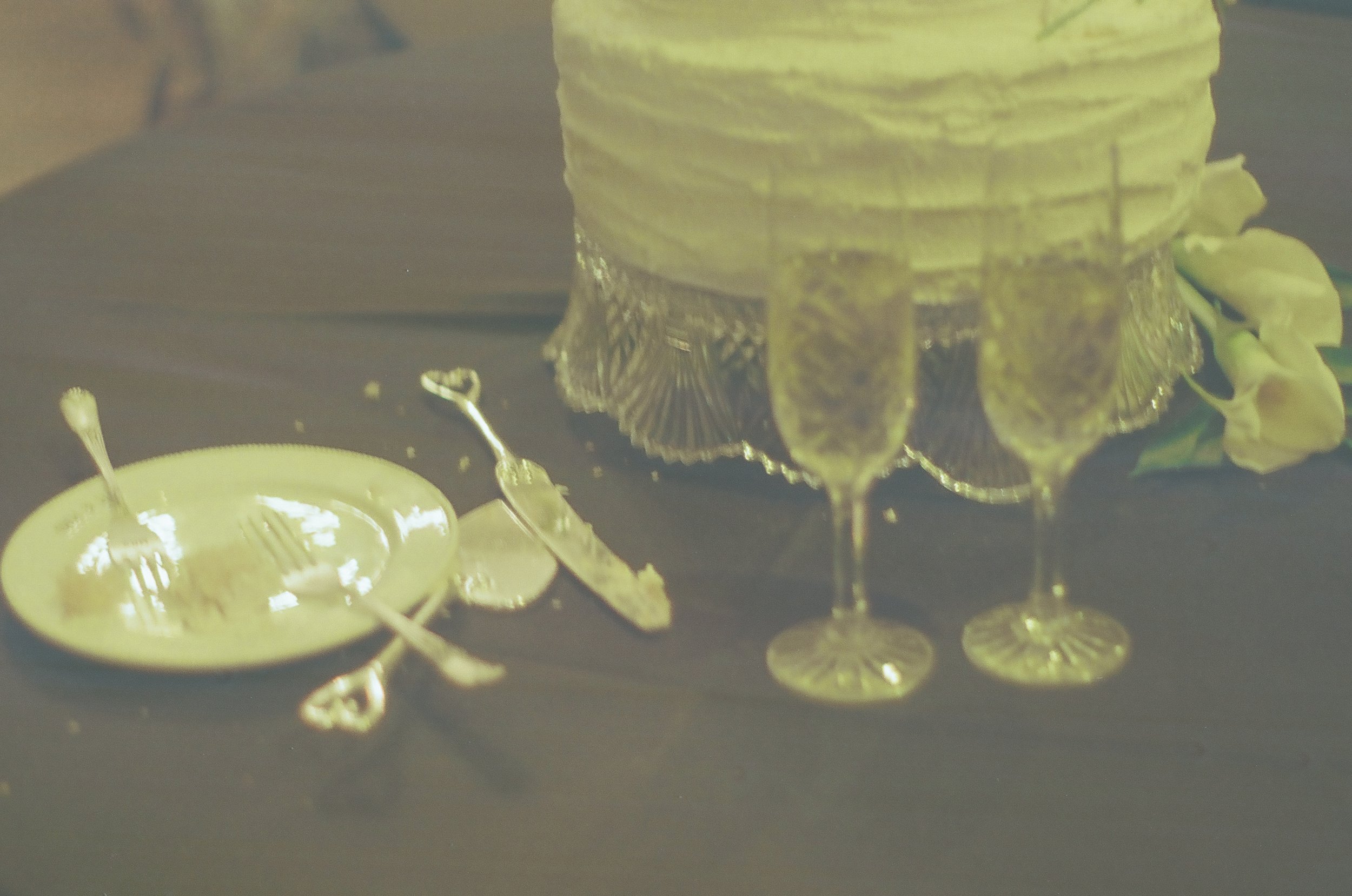 Gold-colored cake, two gold champagne glasses, a gold tray with forks, and a gold flower on a dark table on 35mm film