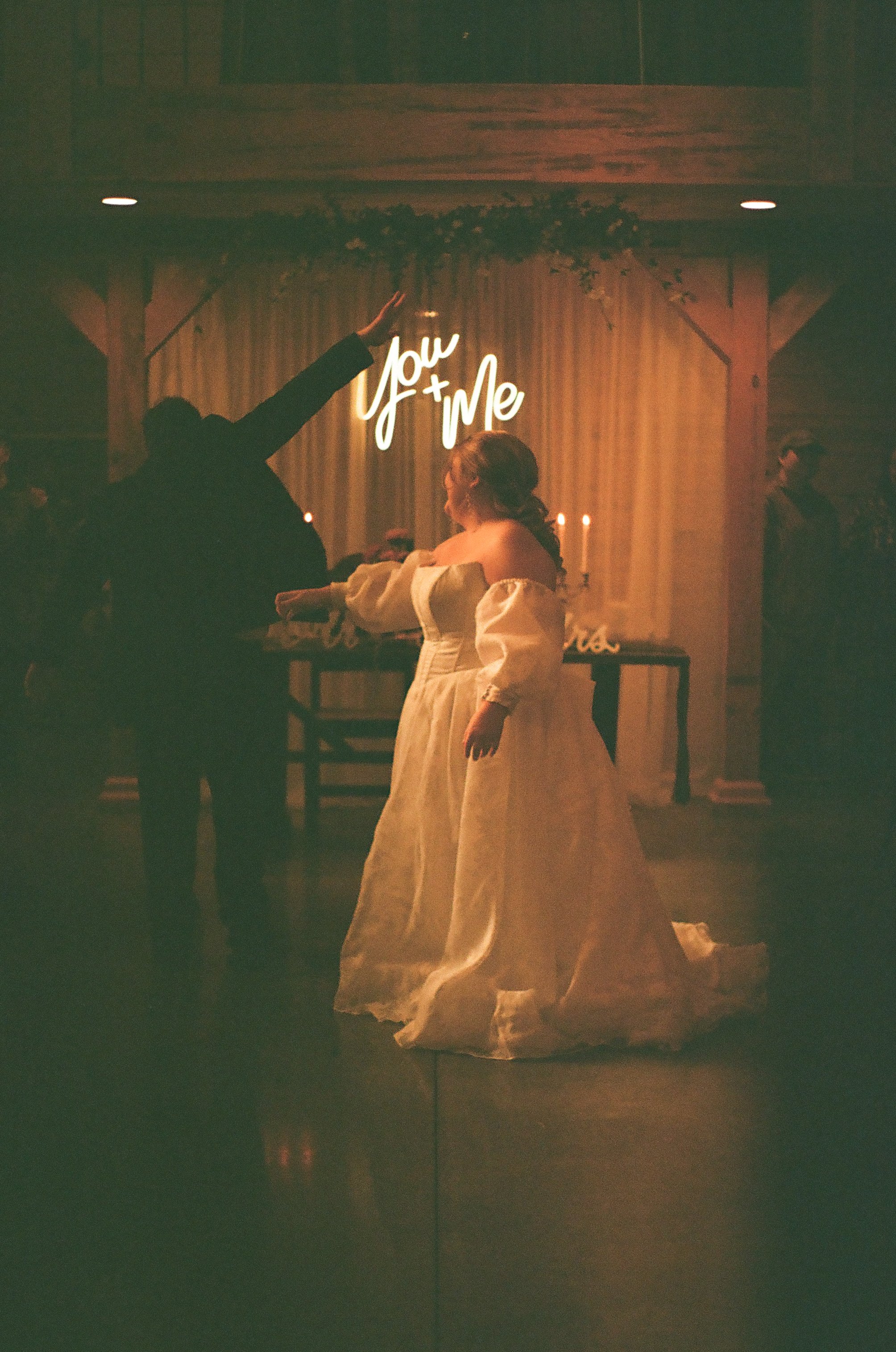 A woman in a white wedding dress at a wedding reception, with a neon sign that says 'You & Me' behind her, as a man raises his hand in a toast or dance gesture.