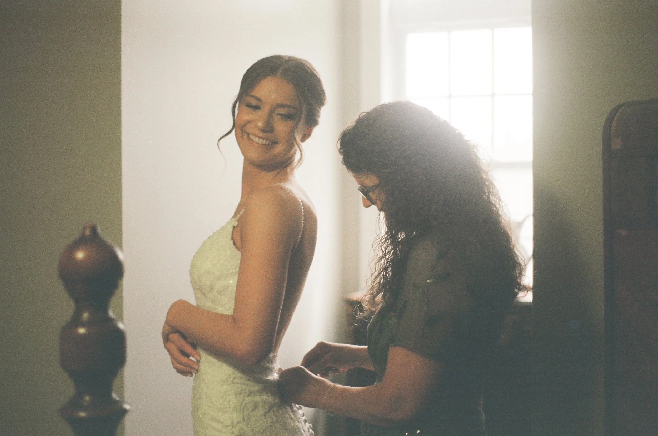 A woman in a wedding dress smiling as a woman helps her with the gown near a window with soft sunlight streaming in.