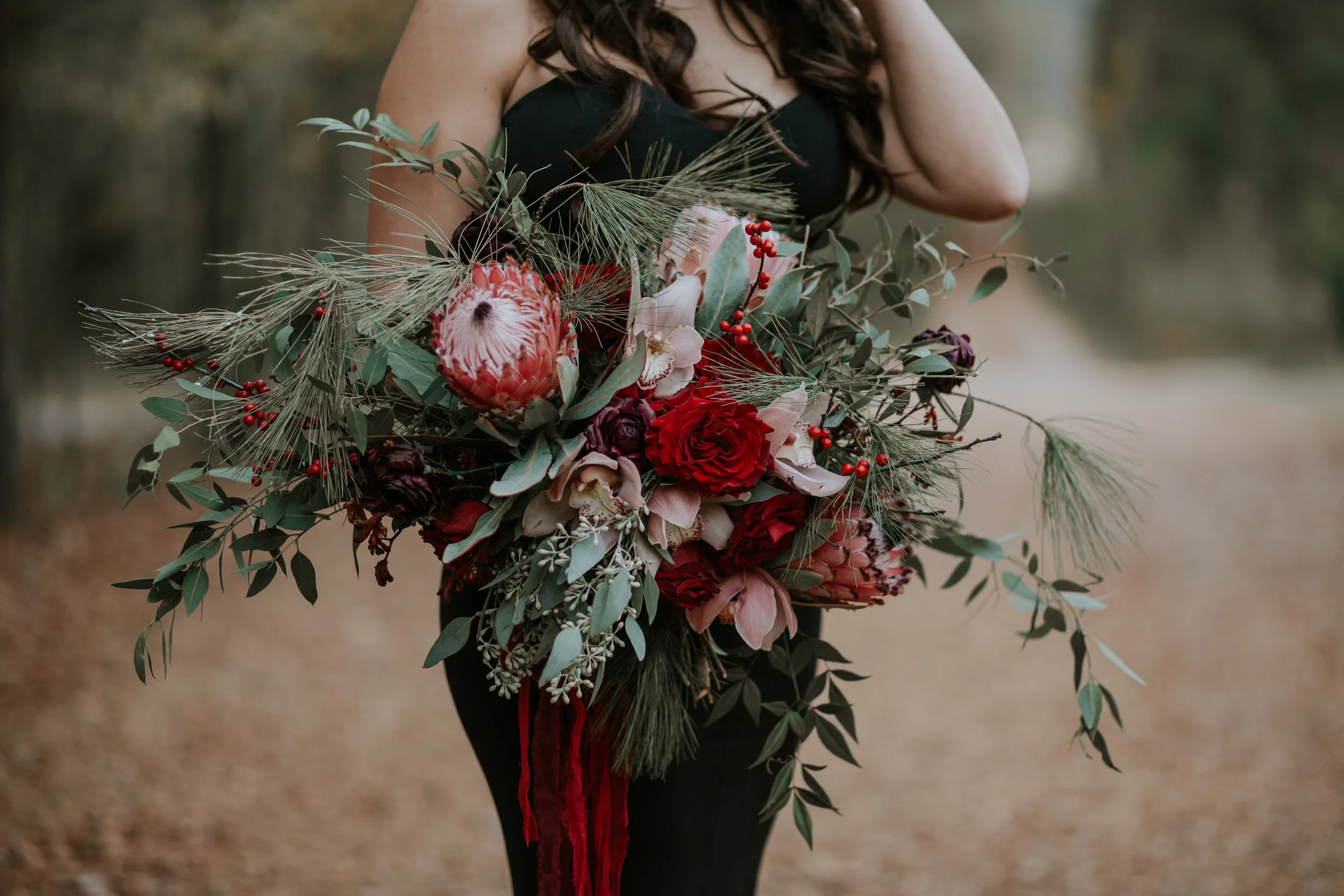 Person wearing a black dress holding a large, abundant bouquet of red roses, pink lilies, protea, green foliage, and berries outdoors.