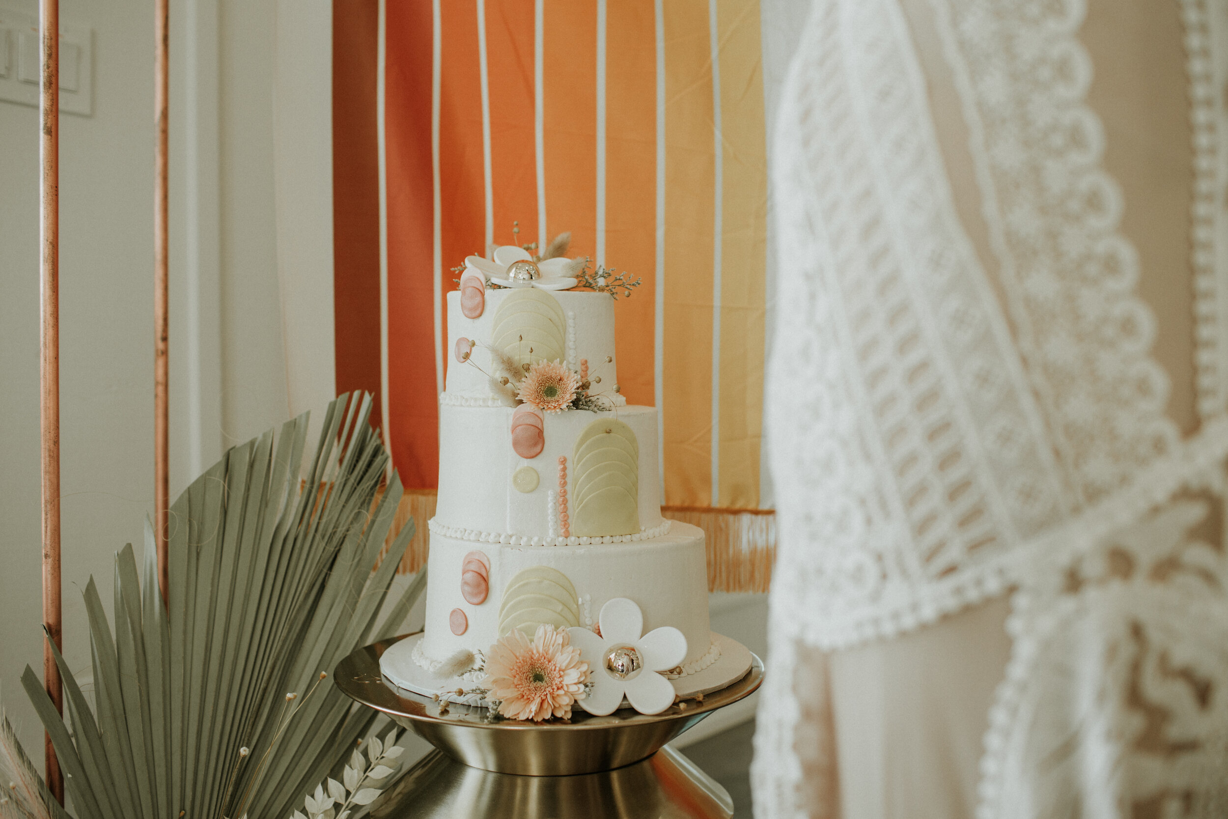 A three-tiered white wedding cake decorated with pink and white flowers, silver accents, and abstract fondant shapes, on a metallic cake stand beside green palm leaves and a lace curtain.