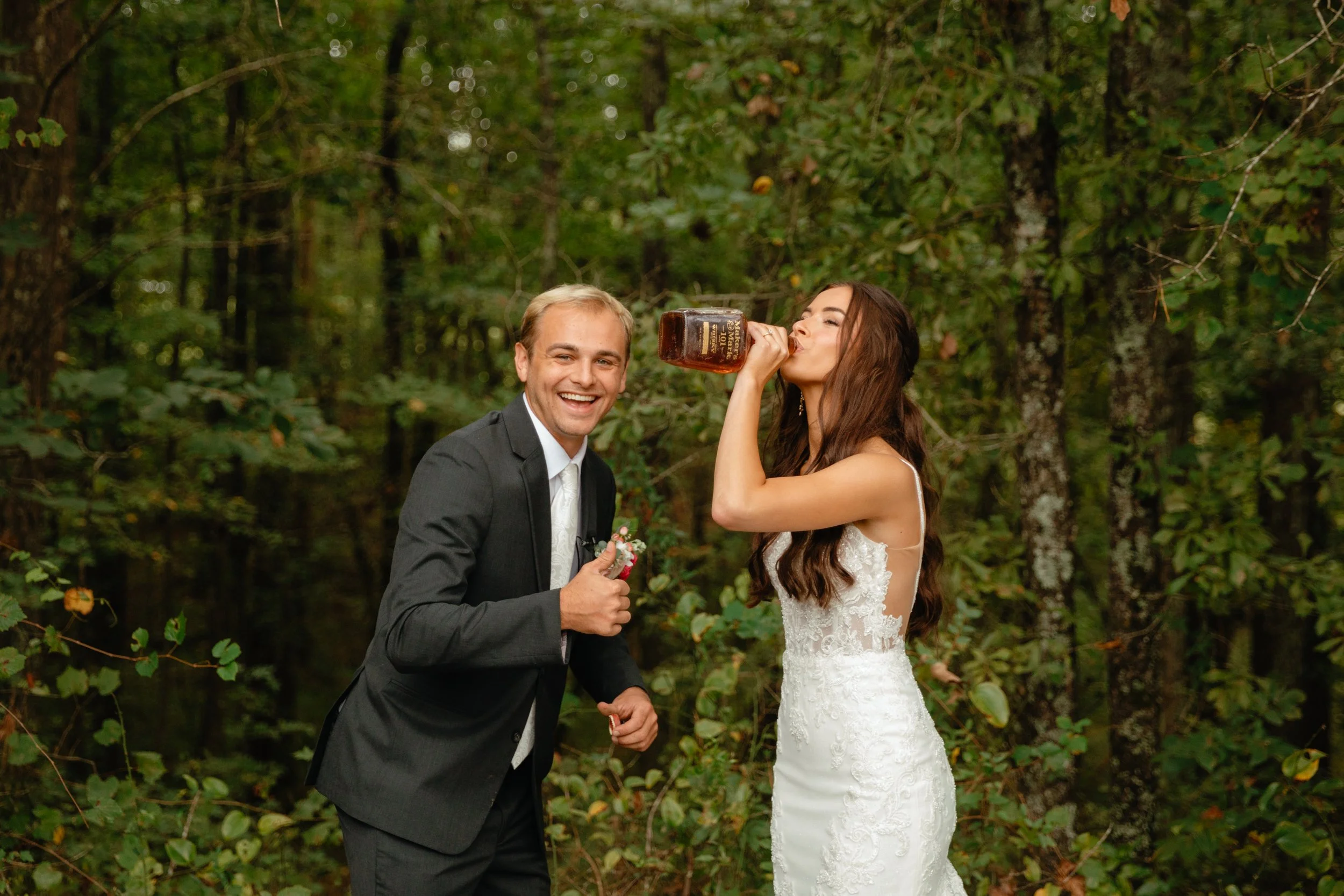 A bride and groom in wedding attire, standing in a forest, with the bride drinking from a bottle and the groom smiling.