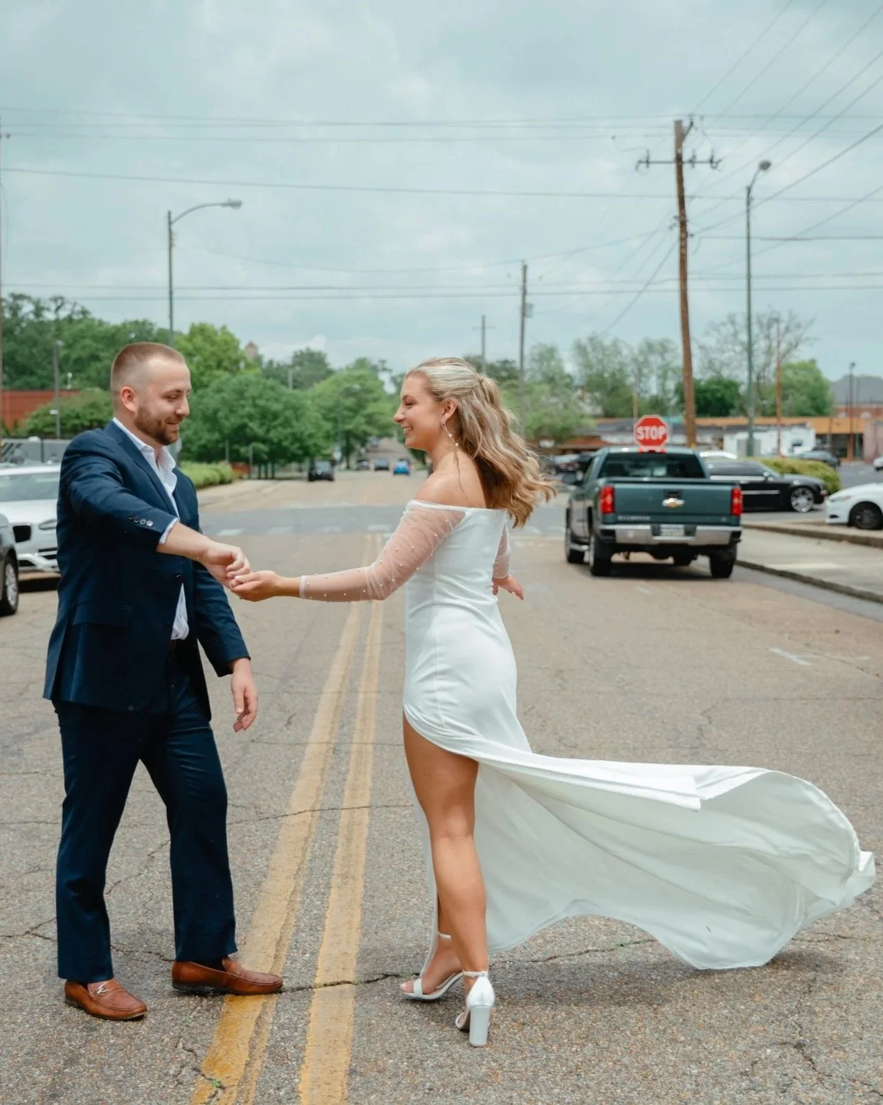A couple dressed in wedding attire on an outdoor street, holding hands and smiling at each other, with the woman's dress flowing behind her.