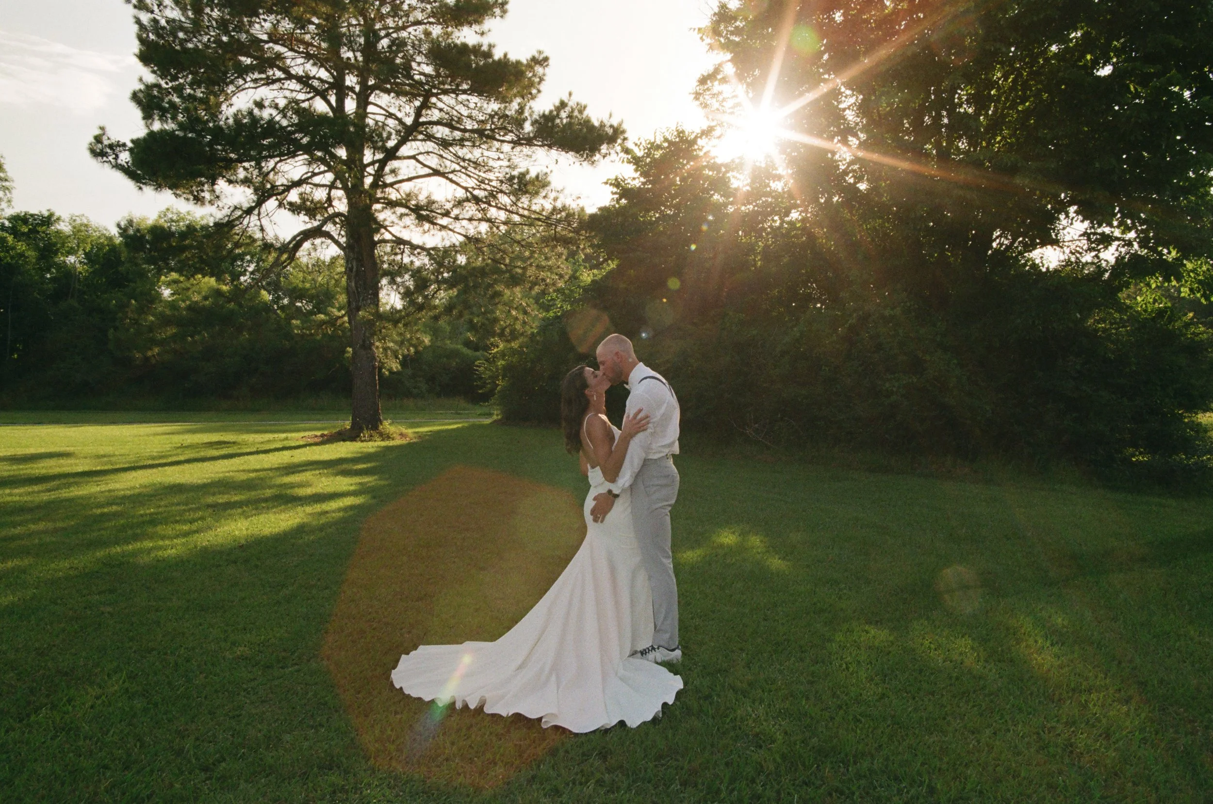 A couple in wedding attire sharing a kiss on a grassy field with trees and sunlight in the background.