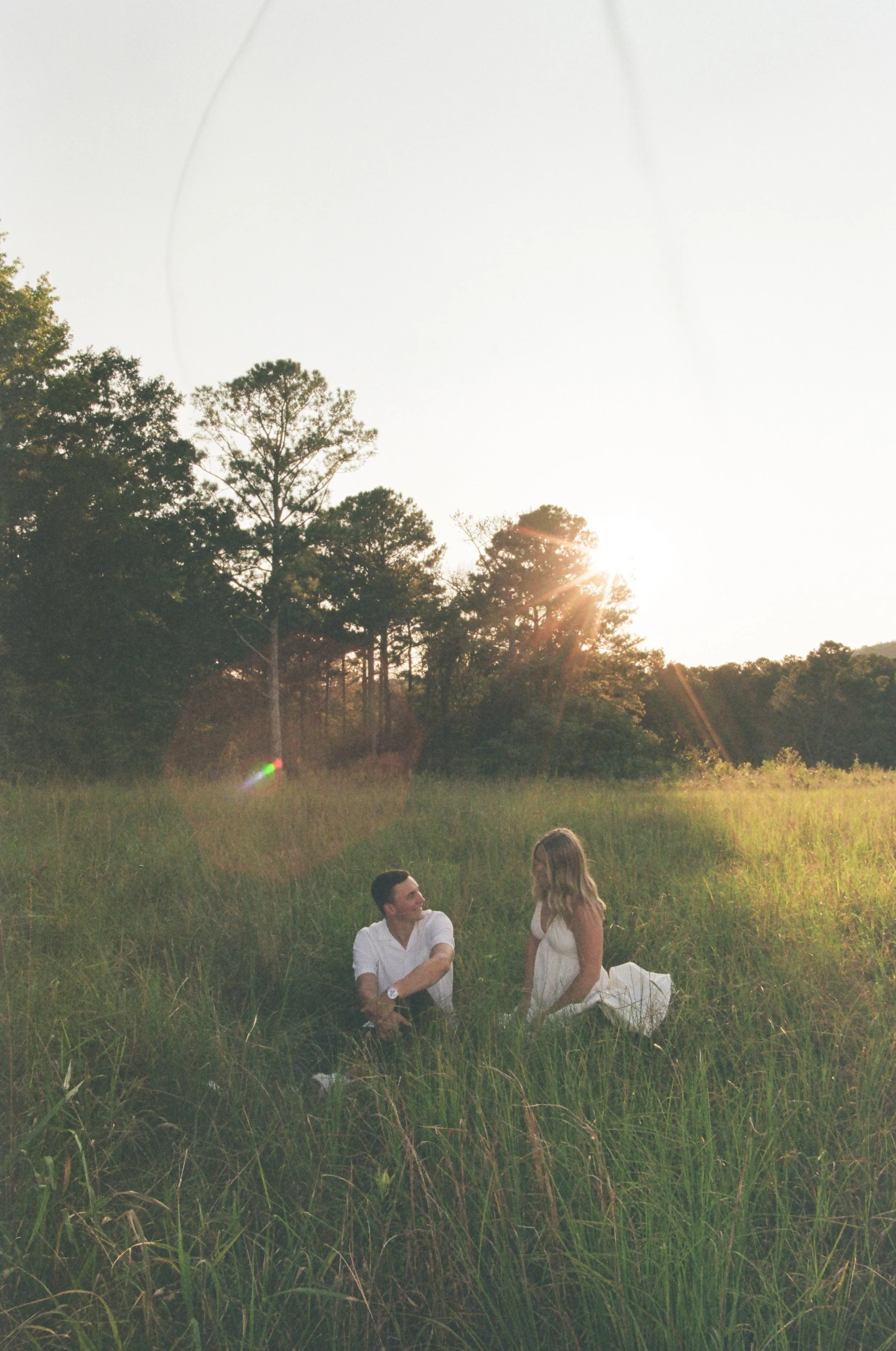 A man and a woman sitting together in a grassy field during sunset, smiling and talking to each other.