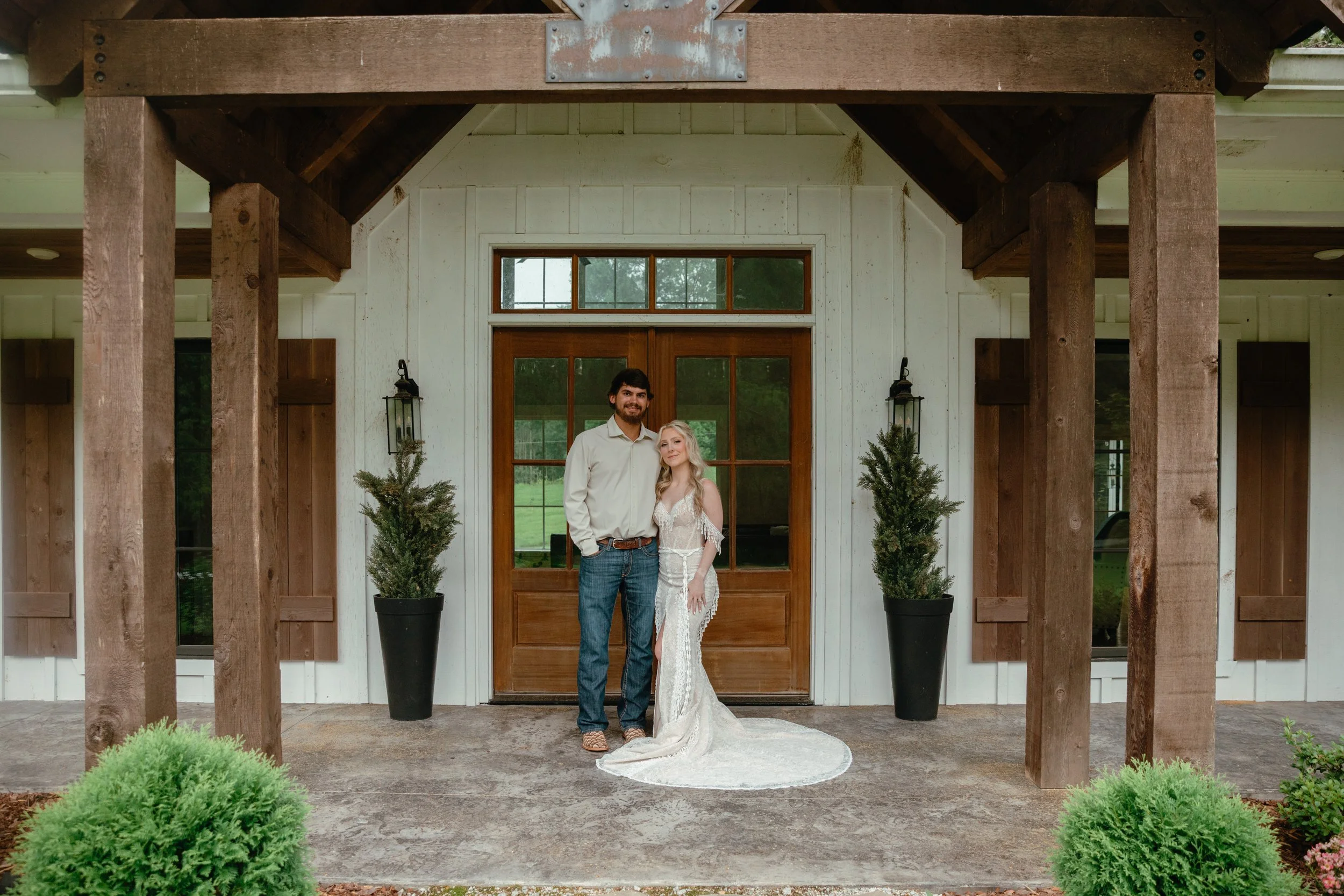 A man and woman stand together in front of a rustic wooden door on a porch, with the man wearing a white shirt and jeans and the woman in a white wedding dress. There are two potted evergreen plants on each side of the door, and a large porch frame w