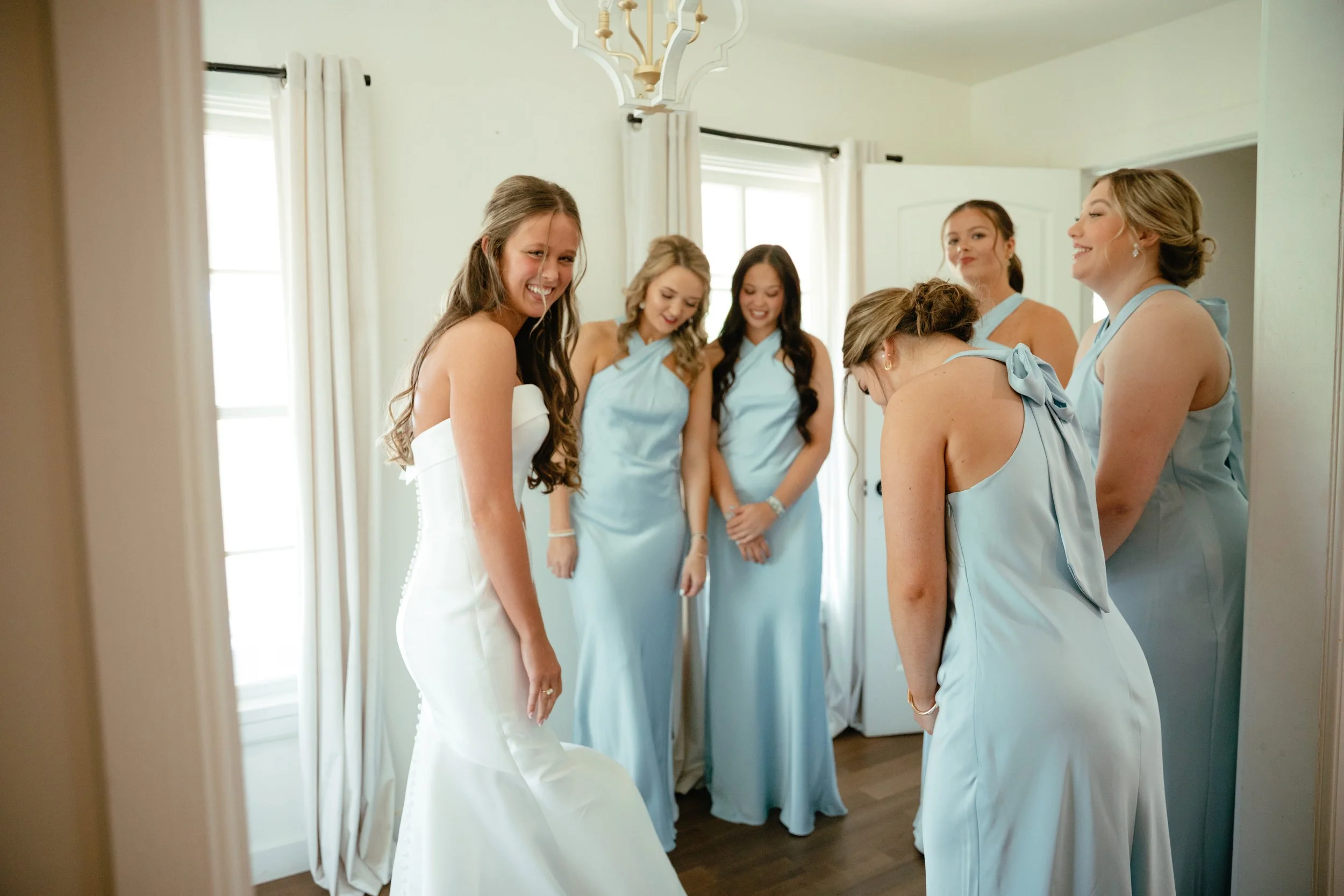 A bride and five bridesmaids preparing before a wedding, smiling and laughing, in a bright room with natural light, with one bridesmaid bowing her head.