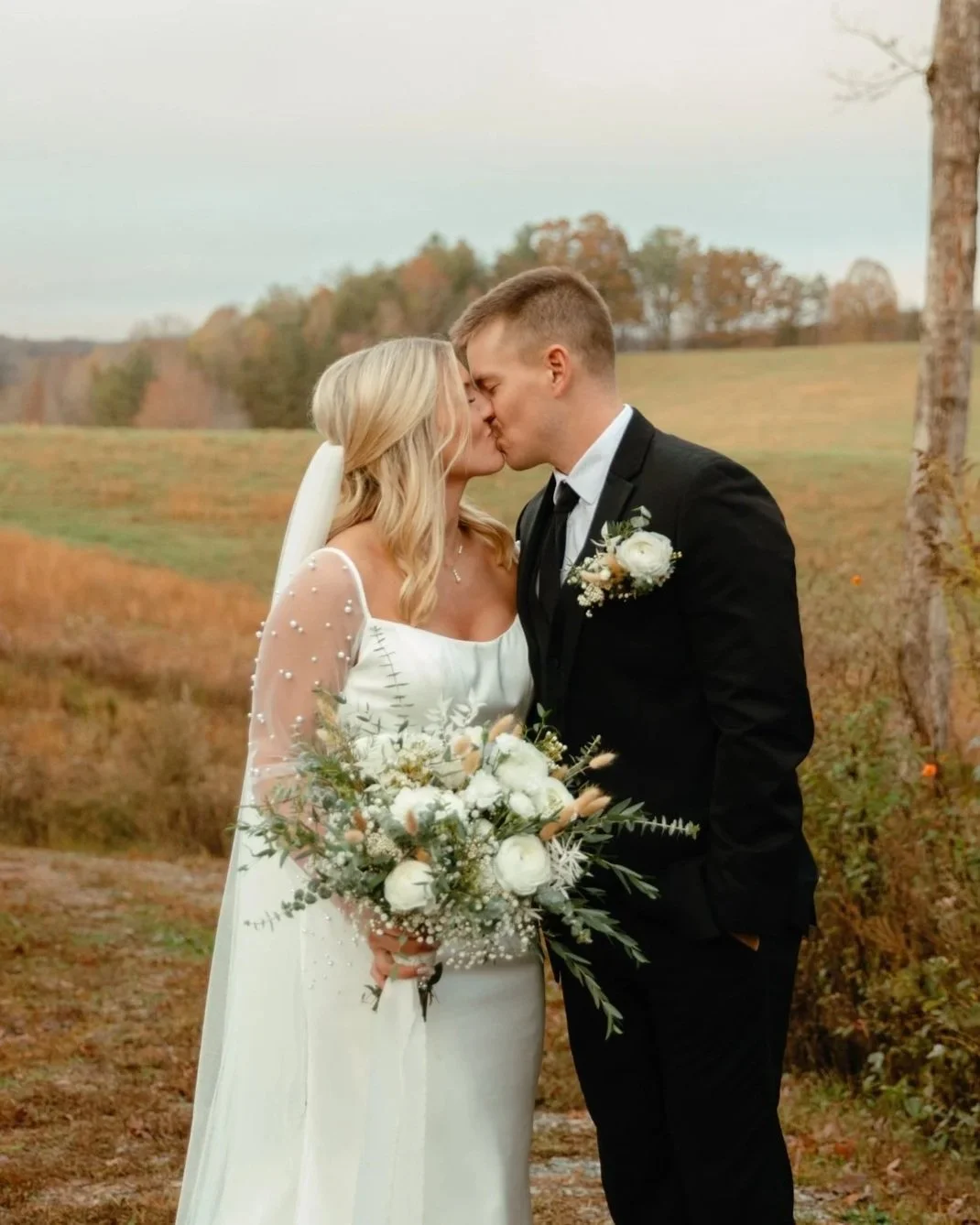 Bride and groom sharing a kiss outdoors, bride holding a bouquet, autumn landscape background.