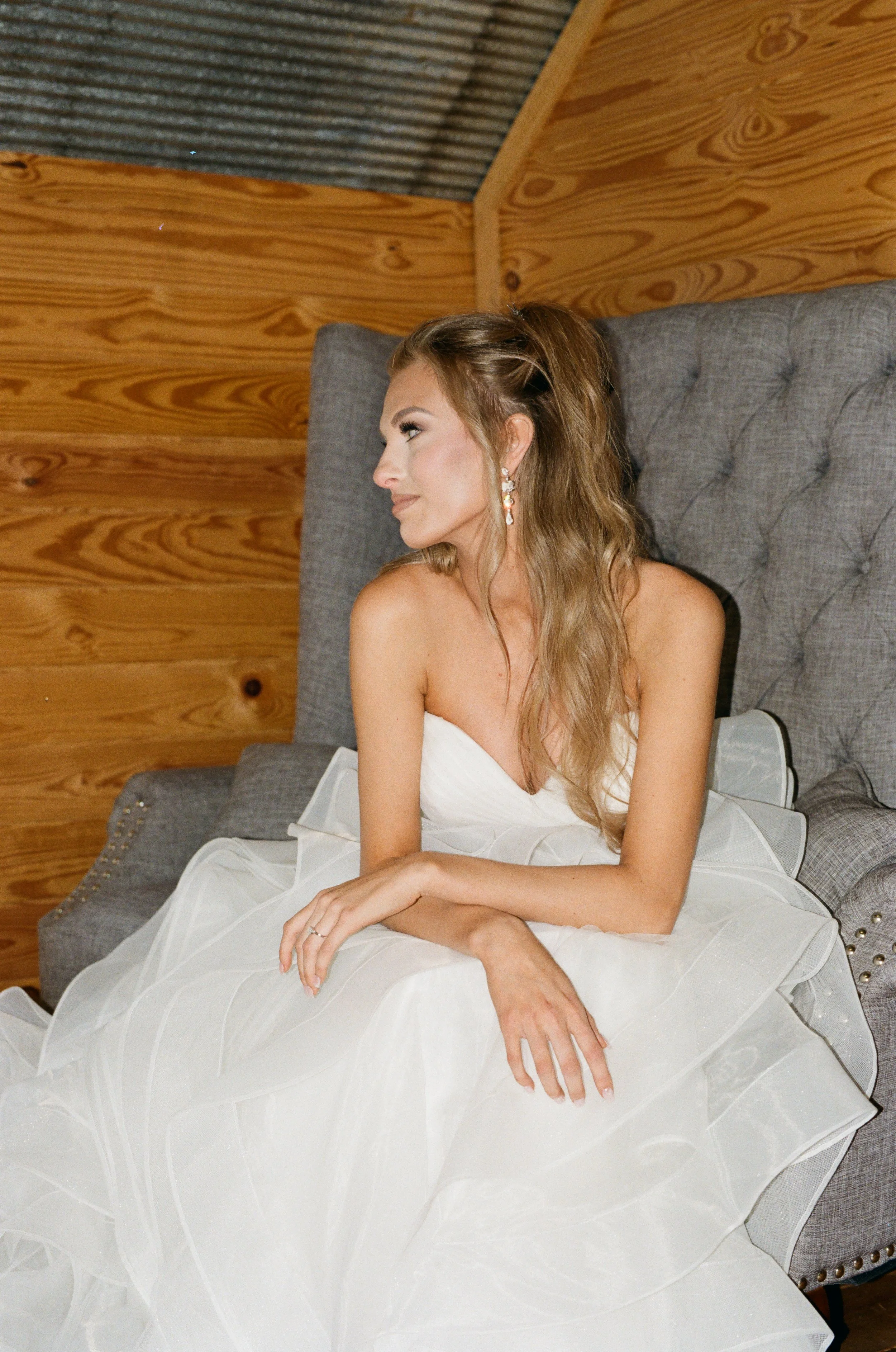 A woman in a white dress sitting on a gray upholstered sofa in a wooden room, looking to the side.  Back 40 farm in jacksonville, Riley Green's venue on 35mm film