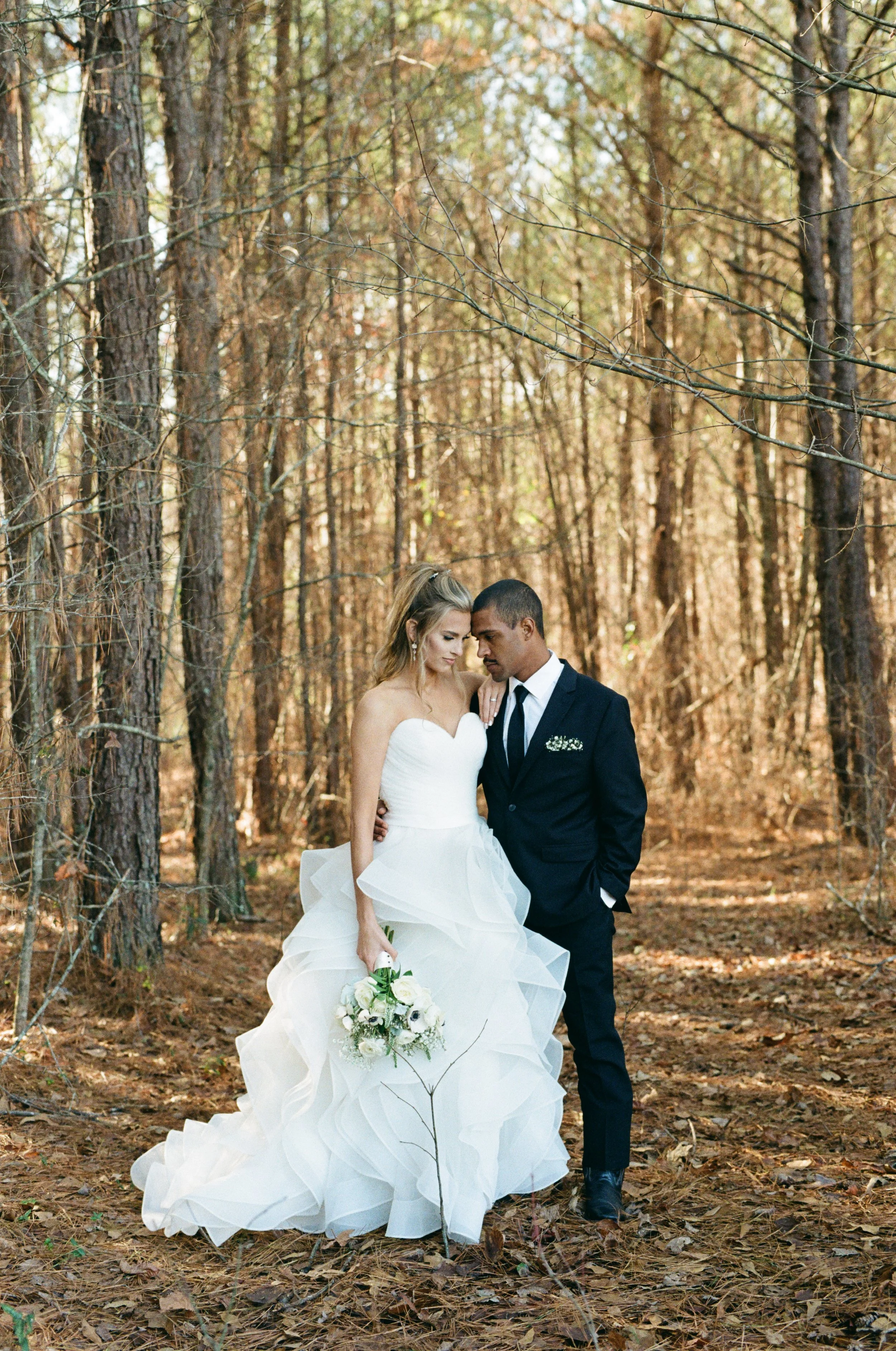 A bride and groom standing close together in a forest, with the bride holding a bouquet of white flowers. The bride wears a strapless white wedding gown with ruffles, and the groom wears a black suit with a white shirt and tie.