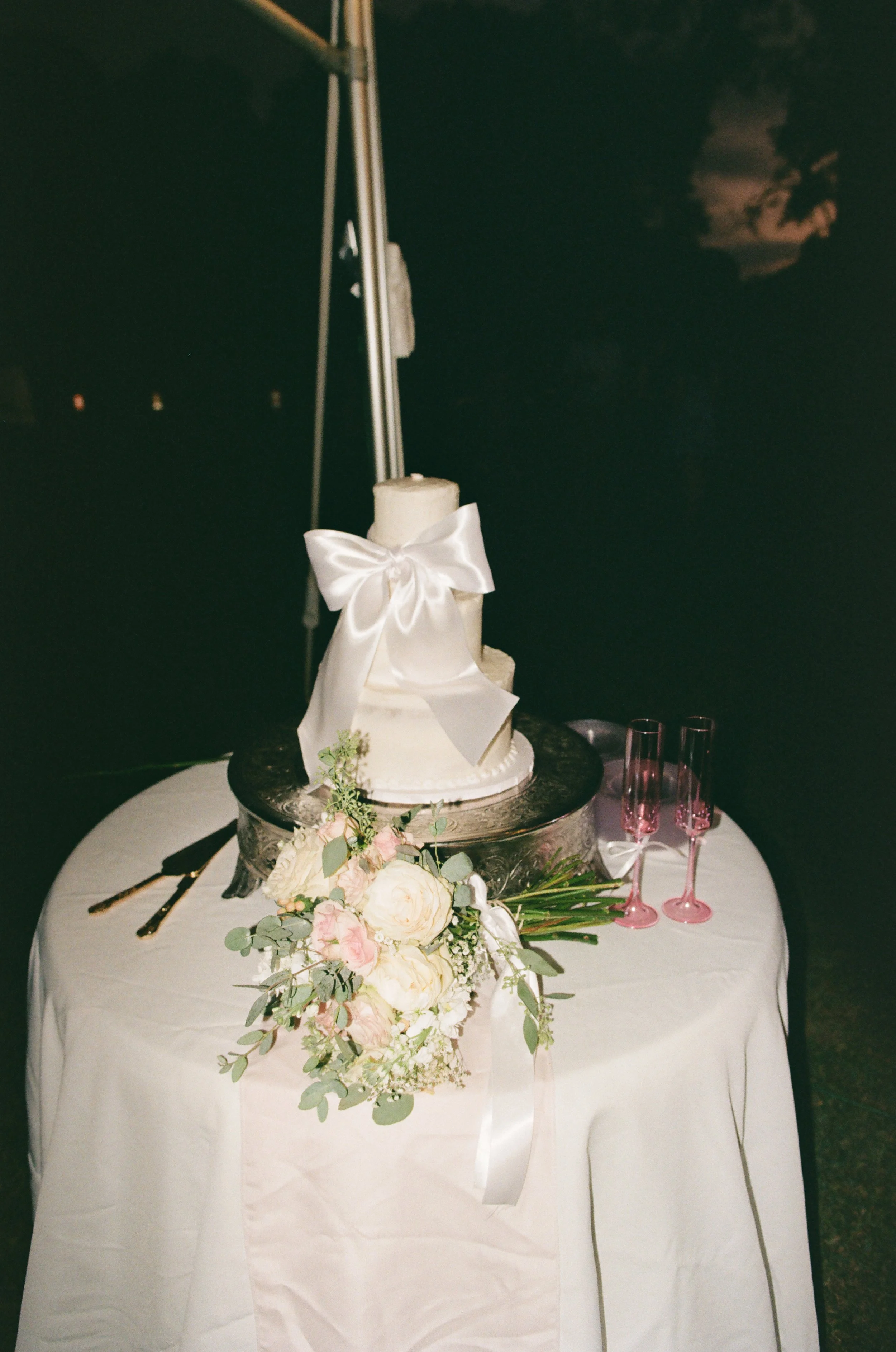A white wedding cake with a satin ribbon bow on top, placed on a silver stand. The cake is decorated with a bouquet of white and light pink flowers and greenery. There are two pink champagne glasses next to the cake on 35mm film