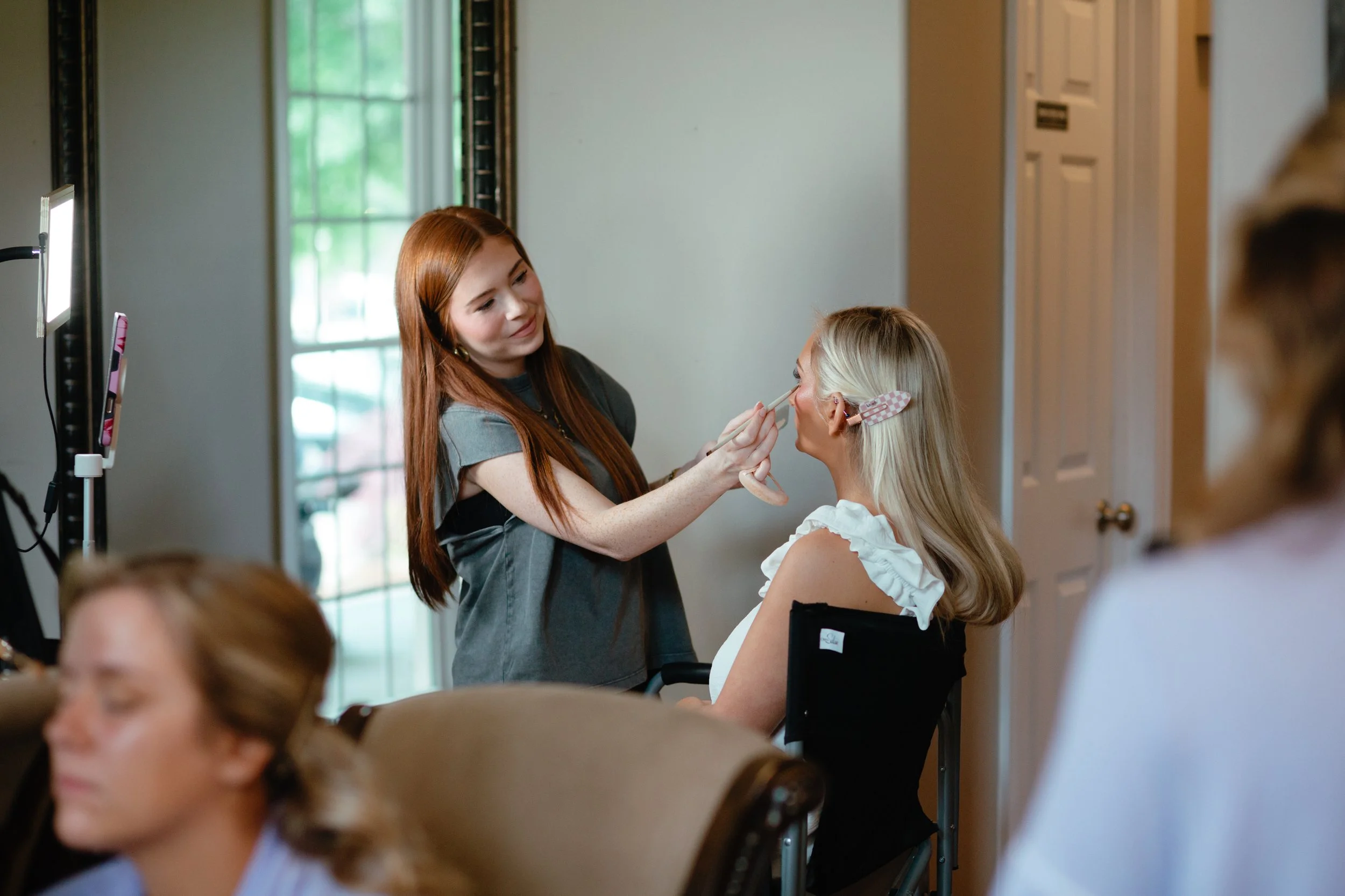 Makeup artist spray-sets a woman's hair in a salon with light wooden walls and large windows in the background.