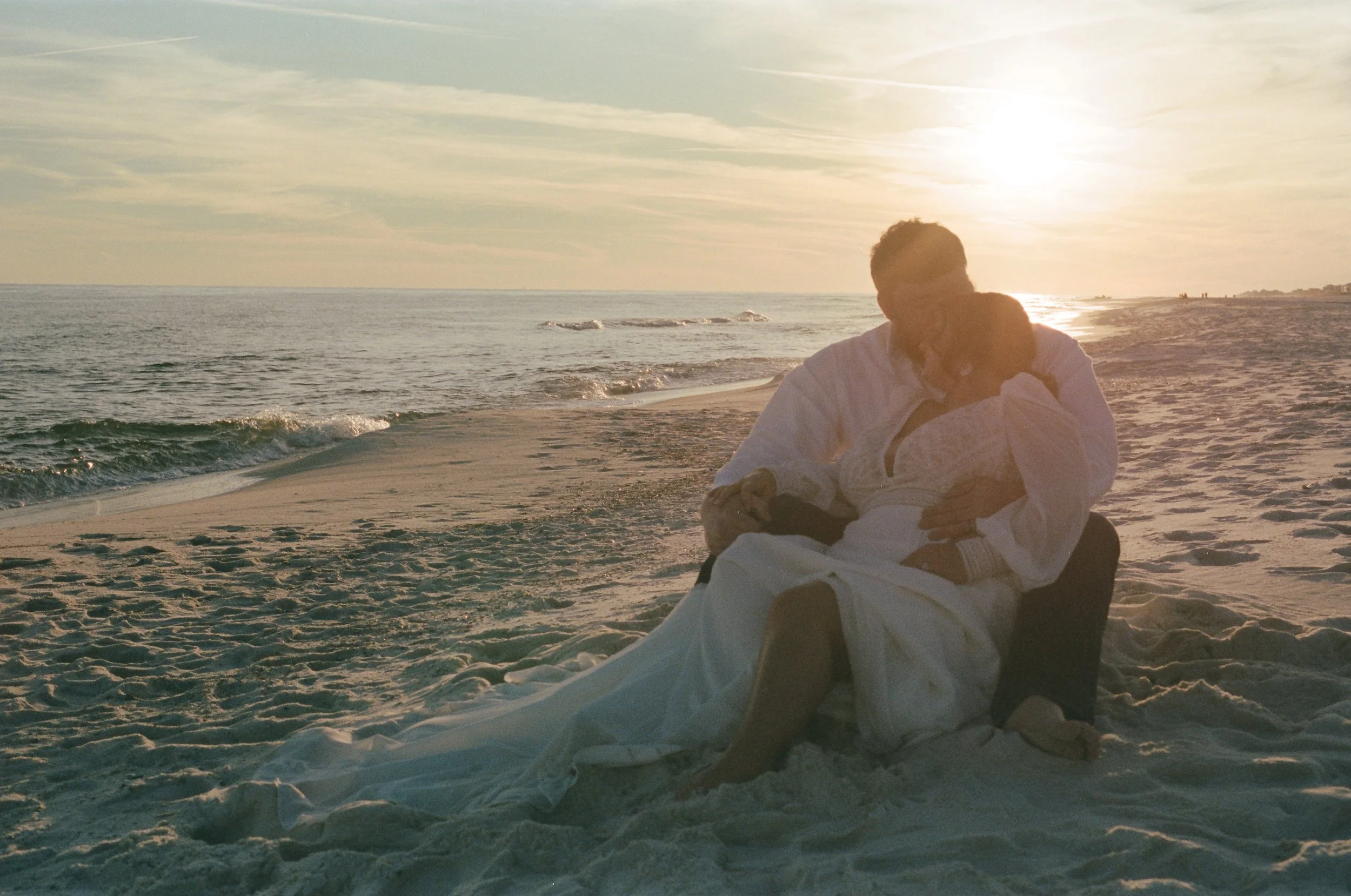 A couple sitting on the beach at sunset, with the ocean and sky in the background. The woman is wearing a white dress, and the man is in a white shirt and dark pants. They are sitting close, embracing and smiling.