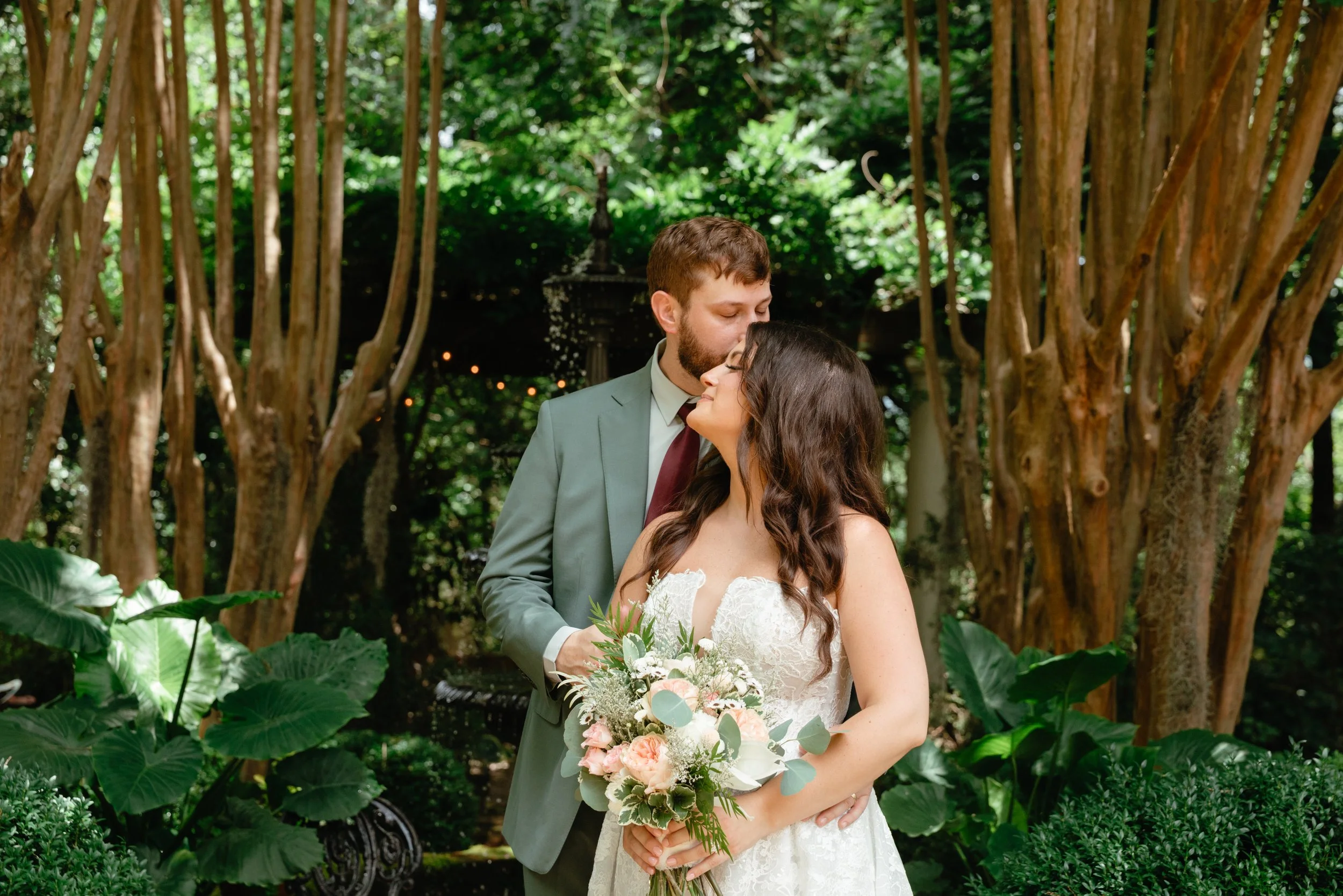 A bride and groom sharing a kiss during their wedding ceremony in a lush, green garden with large leafy plants and tall trees.