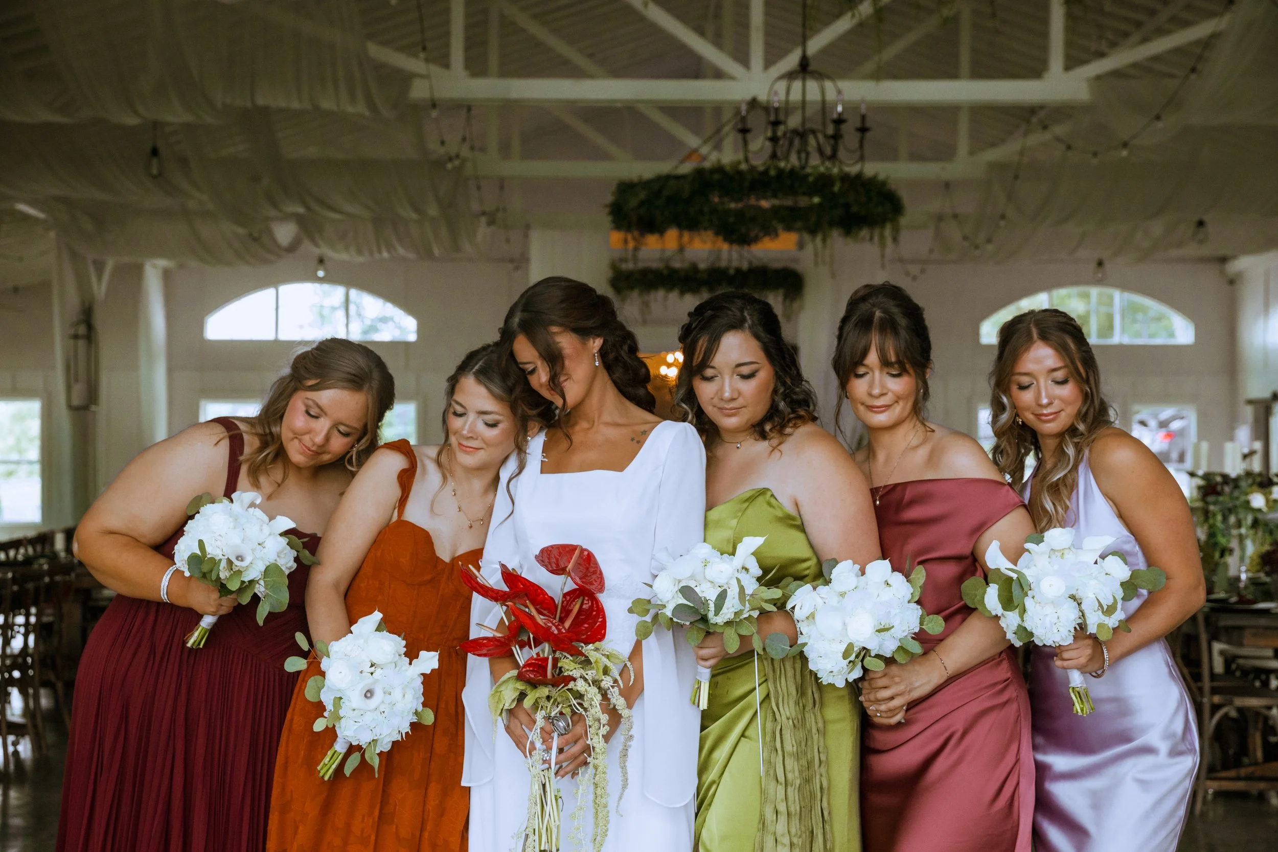 Group of women dressed in colorful gowns holding bouquets, participating in a wedding ceremony inside a decorated venue.