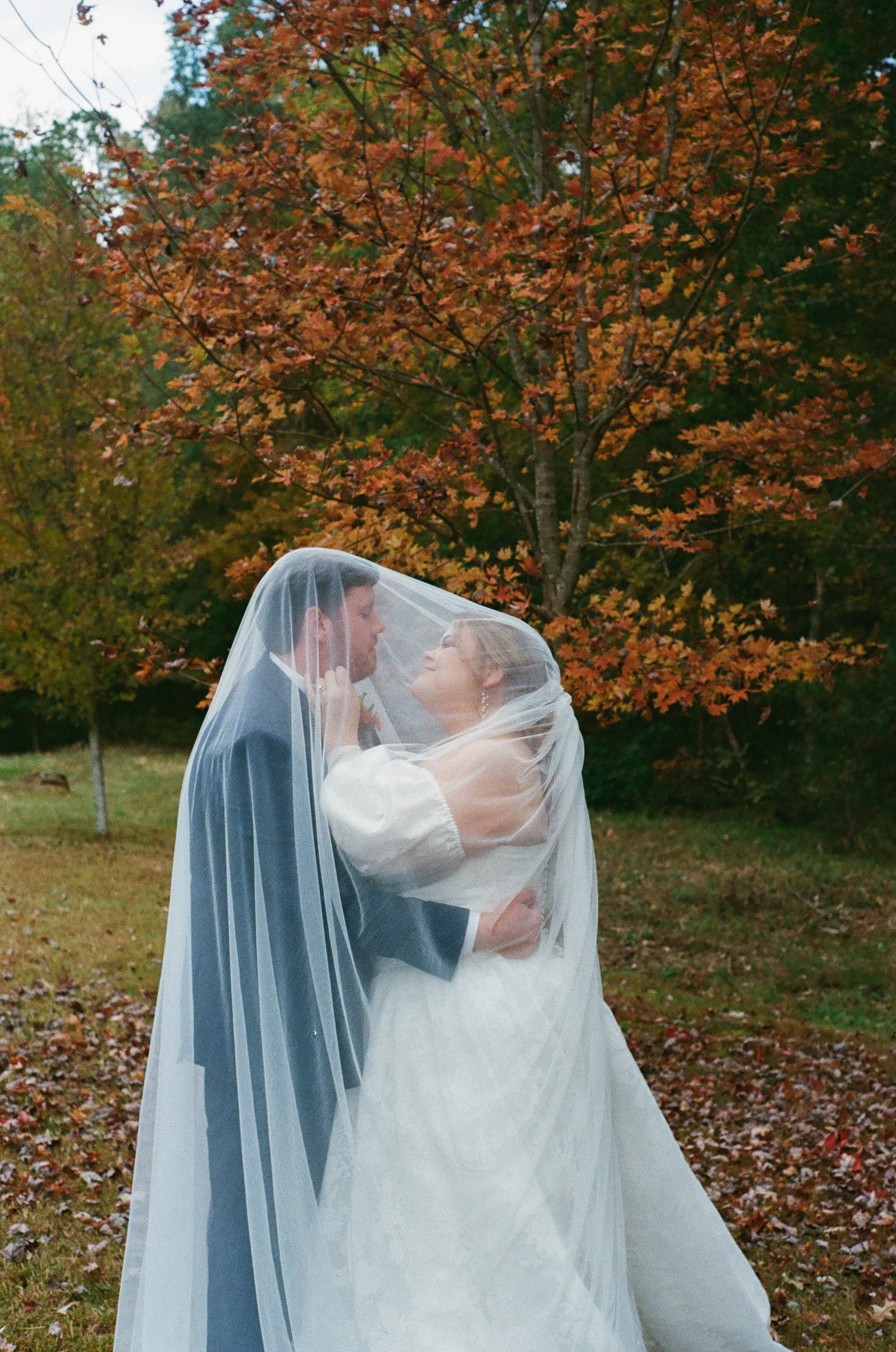 A bride and groom under a wedding veil, standing outdoors in front of autumn-colored trees, with a veil covering both of their faces.