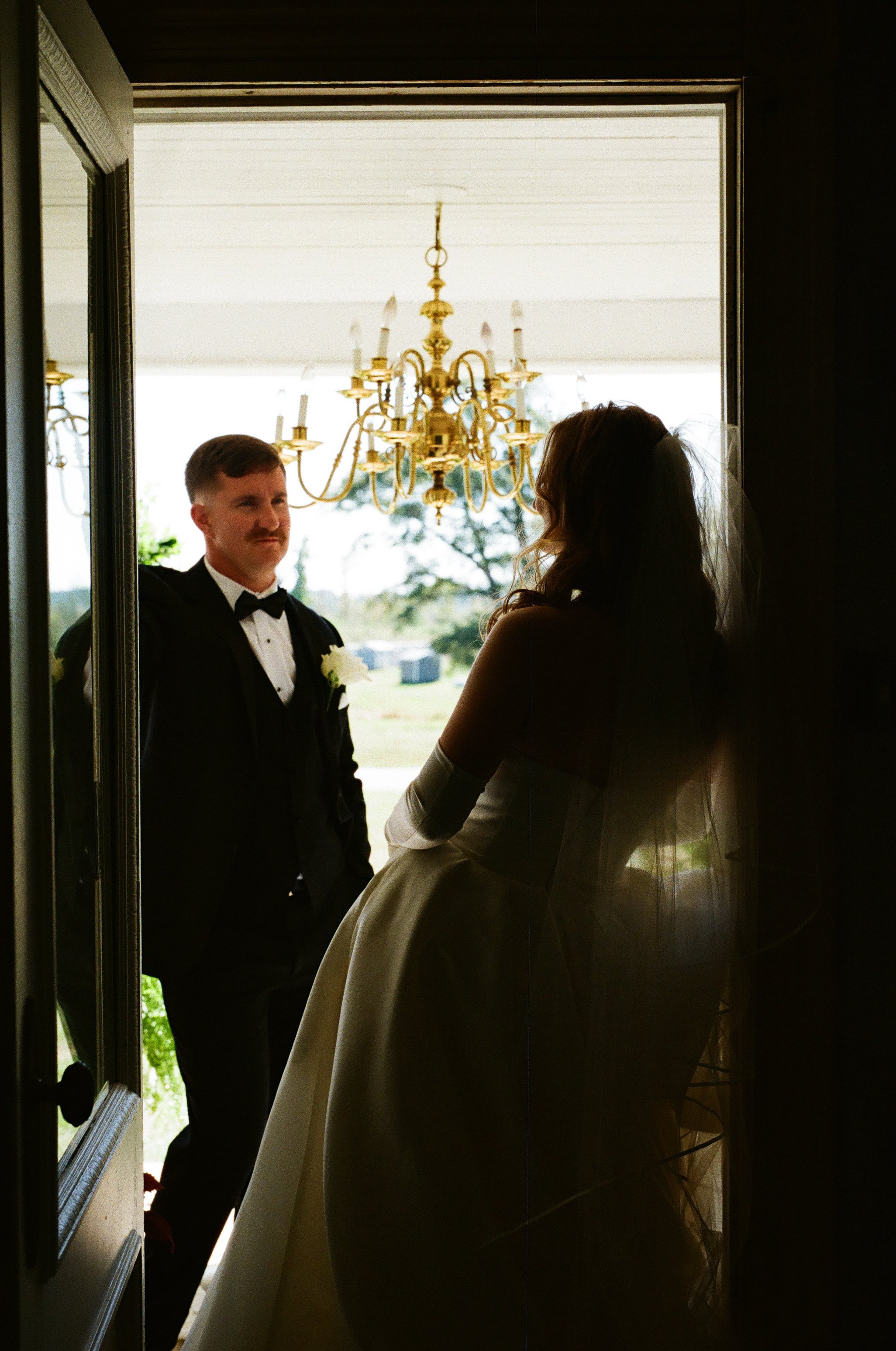 A bride and groom standing in a doorway, with the groom dressed in a black tuxedo and the bride in a wedding gown and veil, backlit by an outdoor scene featuring a chandelier.