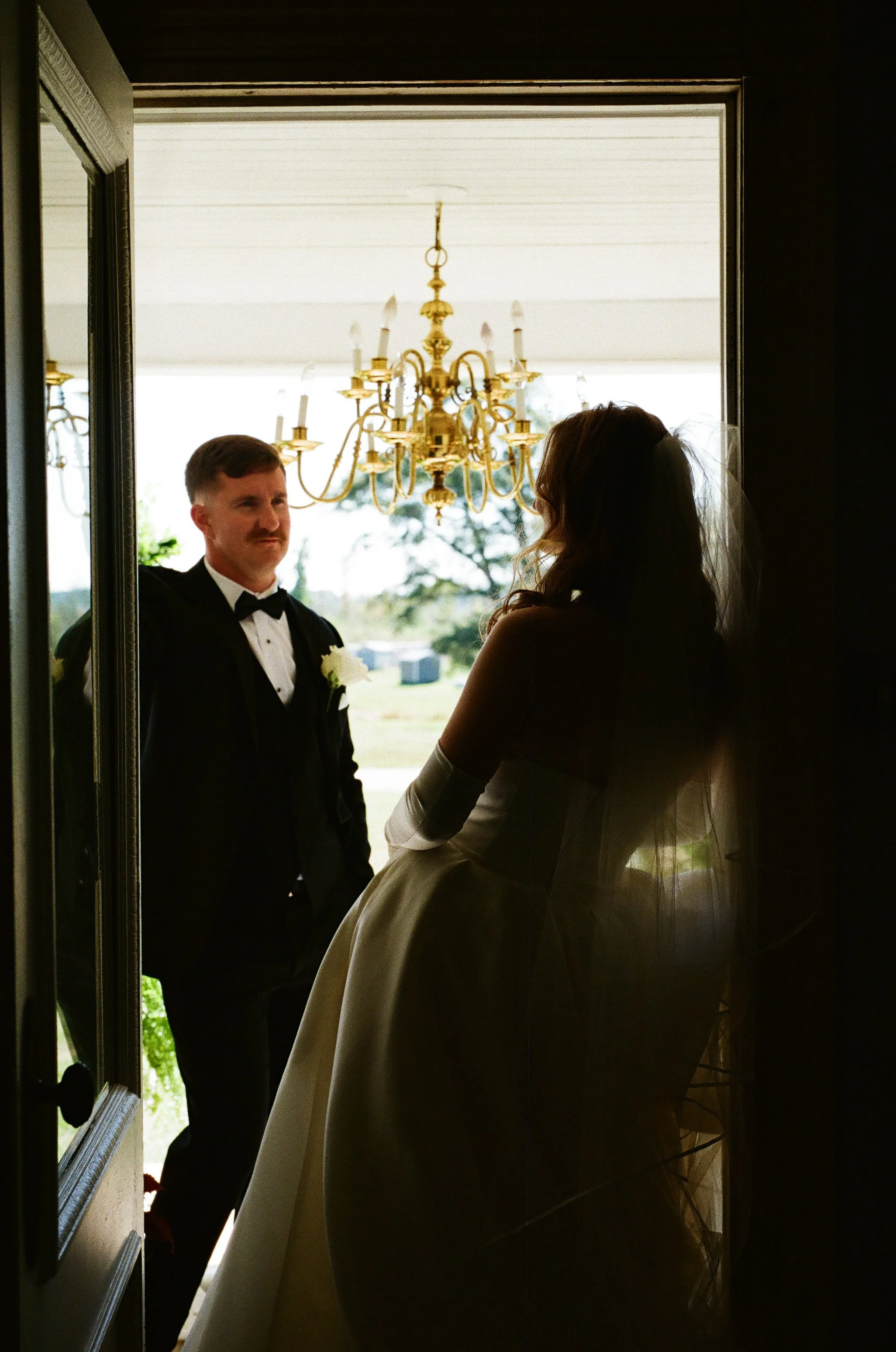 A bride and groom standing in a doorway, with the groom dressed in a black tuxedo and the bride in a wedding gown and veil, backlit by an outdoor scene featuring a chandelier. on 35mm film