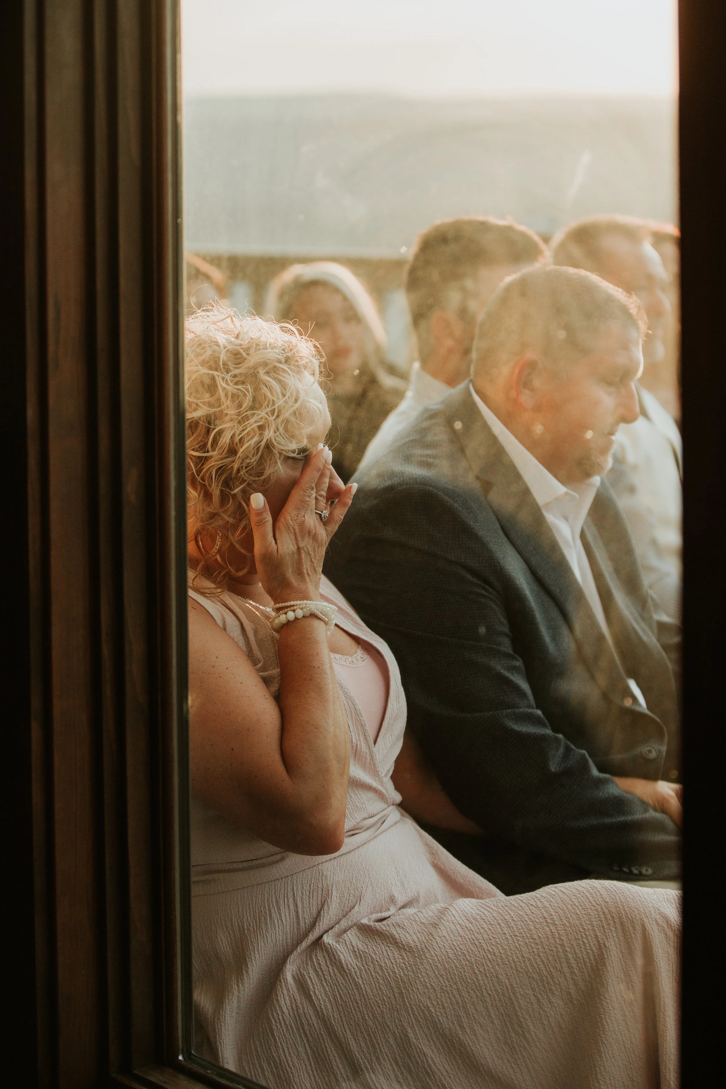 The groom's mother covers her face with her hand, possibly wiping tears, while the man sits next to her. They are viewed through a glass window, with other people visible in the background.