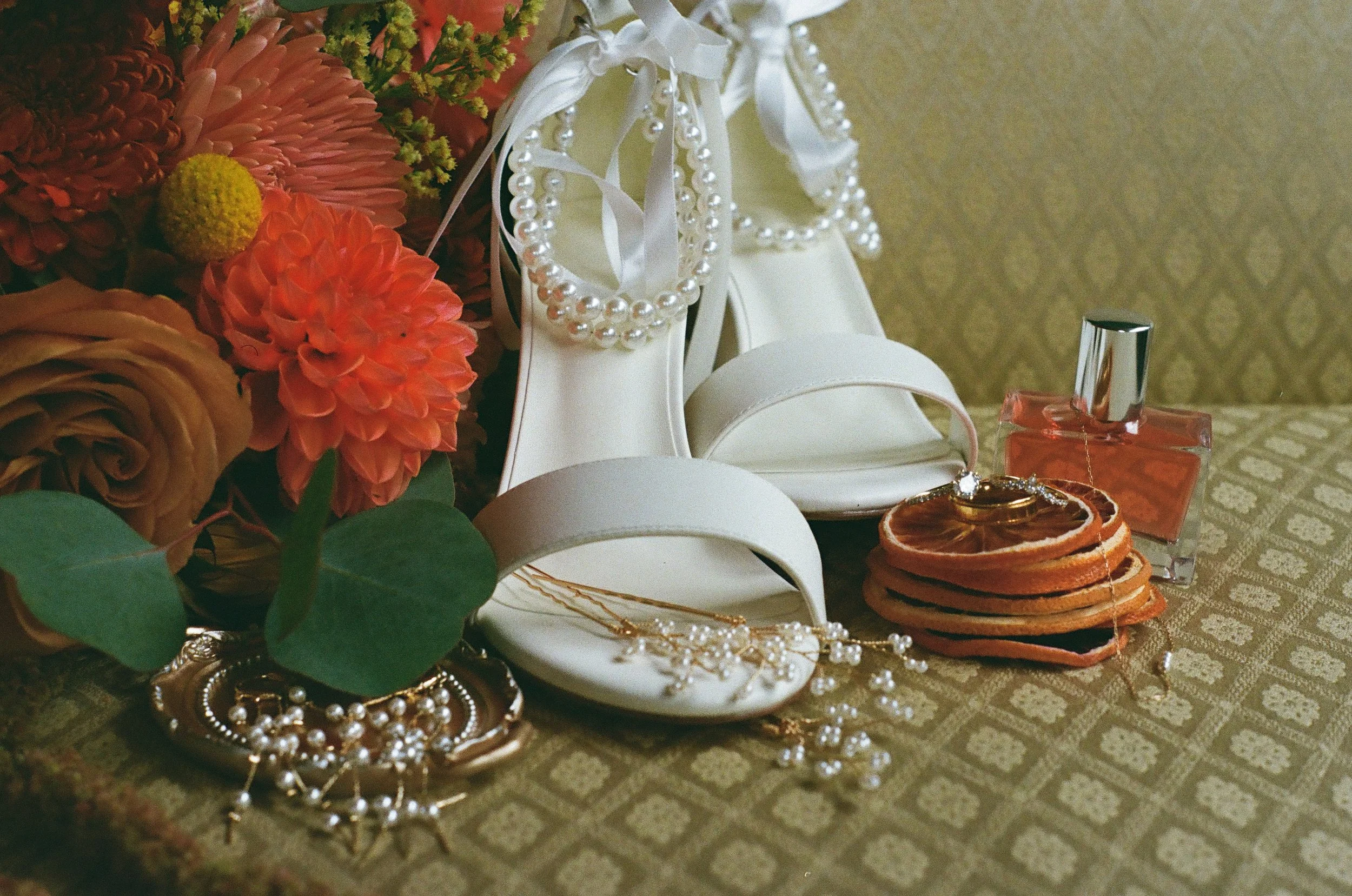 White high-heeled shoes, pearl necklaces, rings, and a bracelet arranged on a table with a floral bouquet, orange slices, and a bottle of pink perfume.