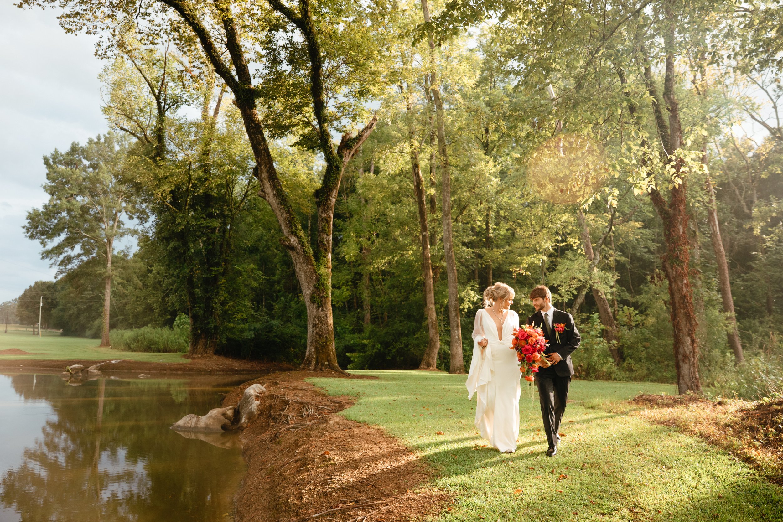 A bride and groom walking along a grassy path beside a small pond in a wooded area during daytime, with sunlight filtering through the trees.