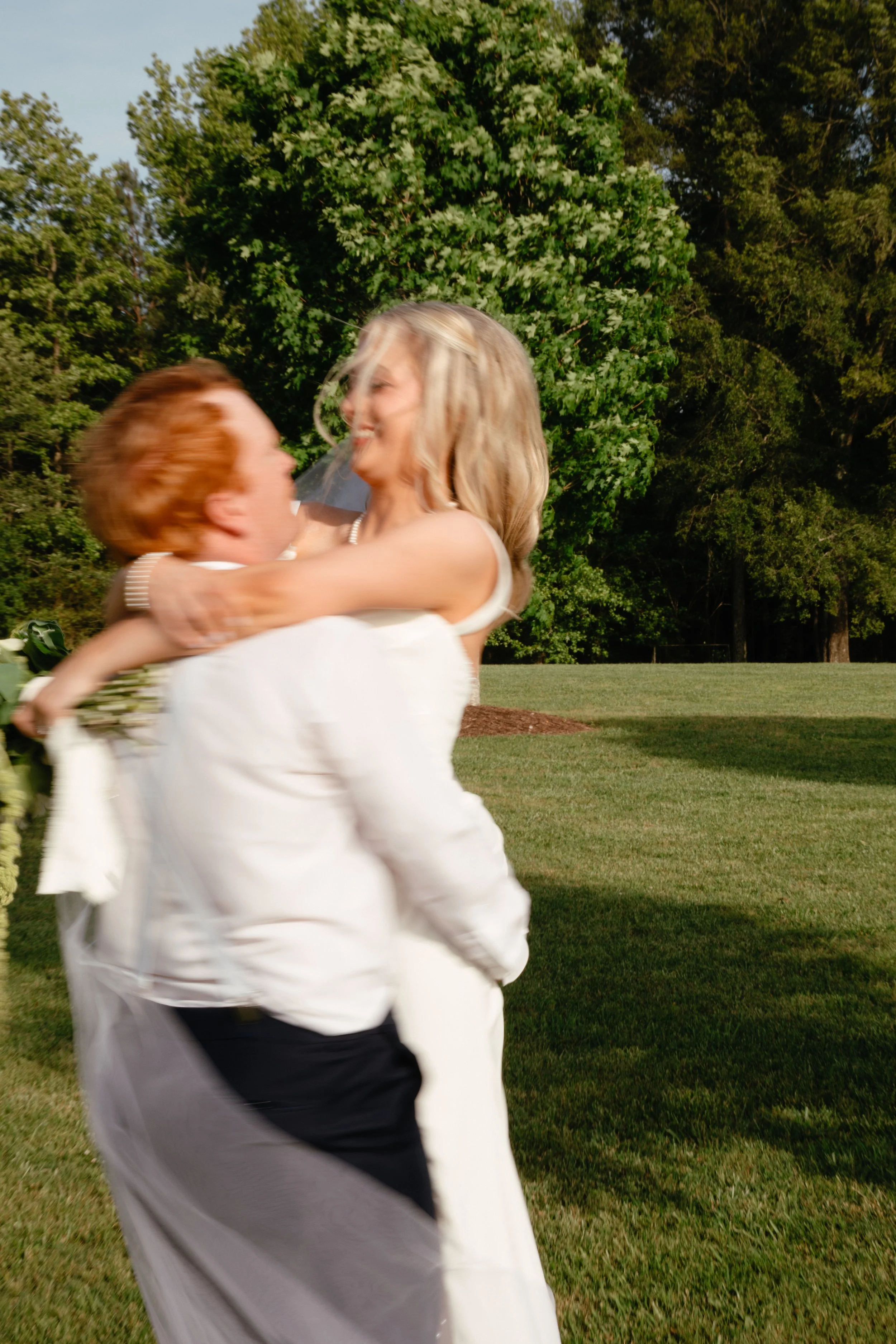A couple, dressed in wedding attire, celebrating outdoors on a grassy field with trees in the background. The woman is being lifted by the man, and they are smiling and embracing each other at The Hay Barn, Birmingham, Alabama.