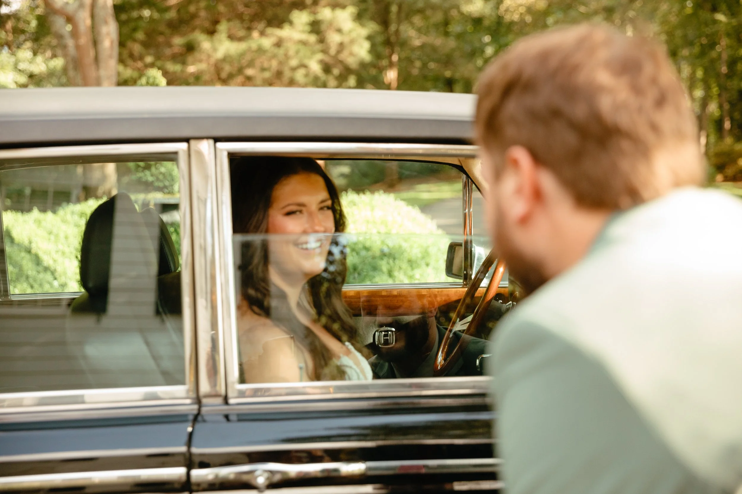 A woman smiling inside a vintage car, with a man partially visible outside the car, in a lush green outdoor setting.