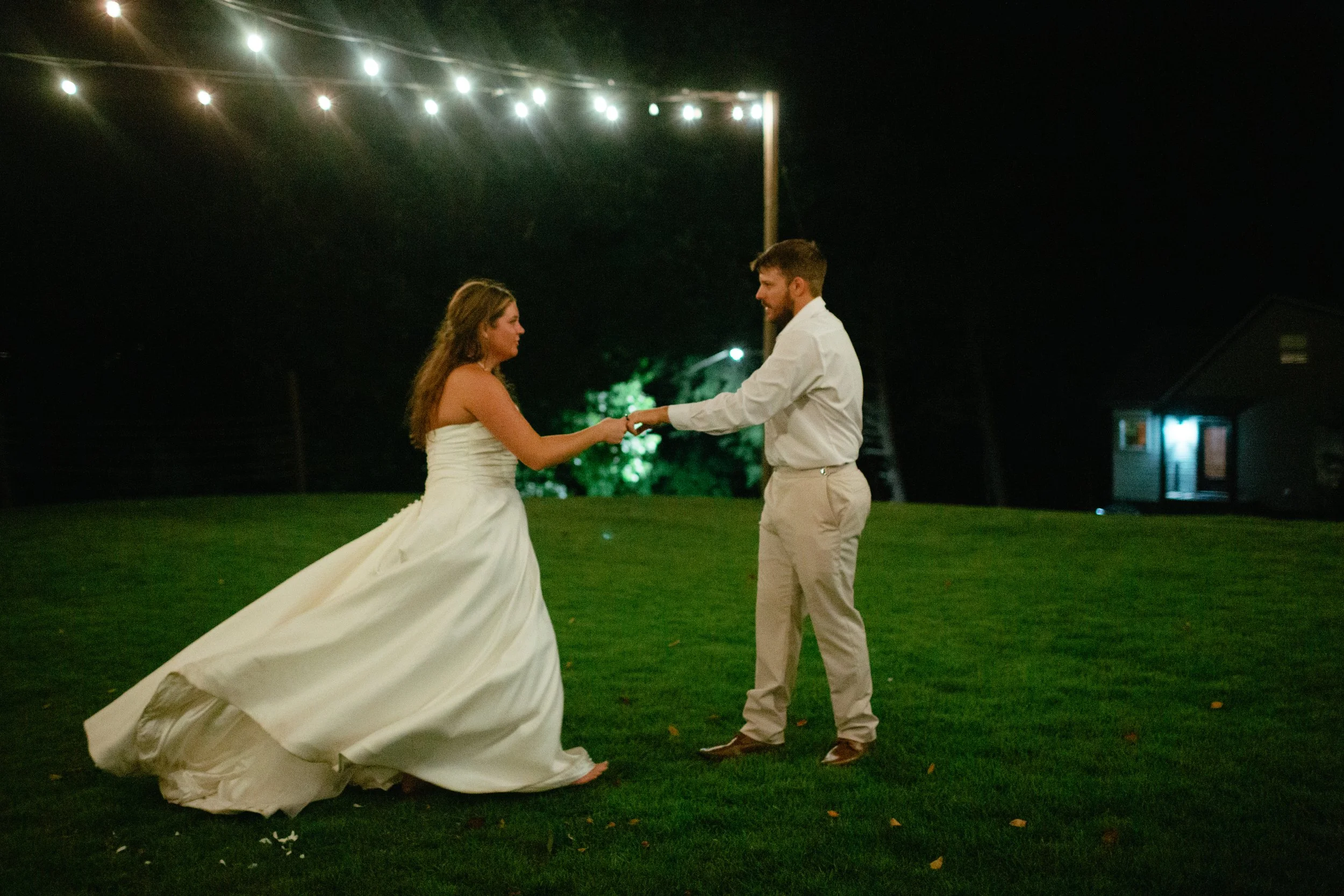 A bride and groom dancing outdoors at night, with string lights overhead and a house in the background.