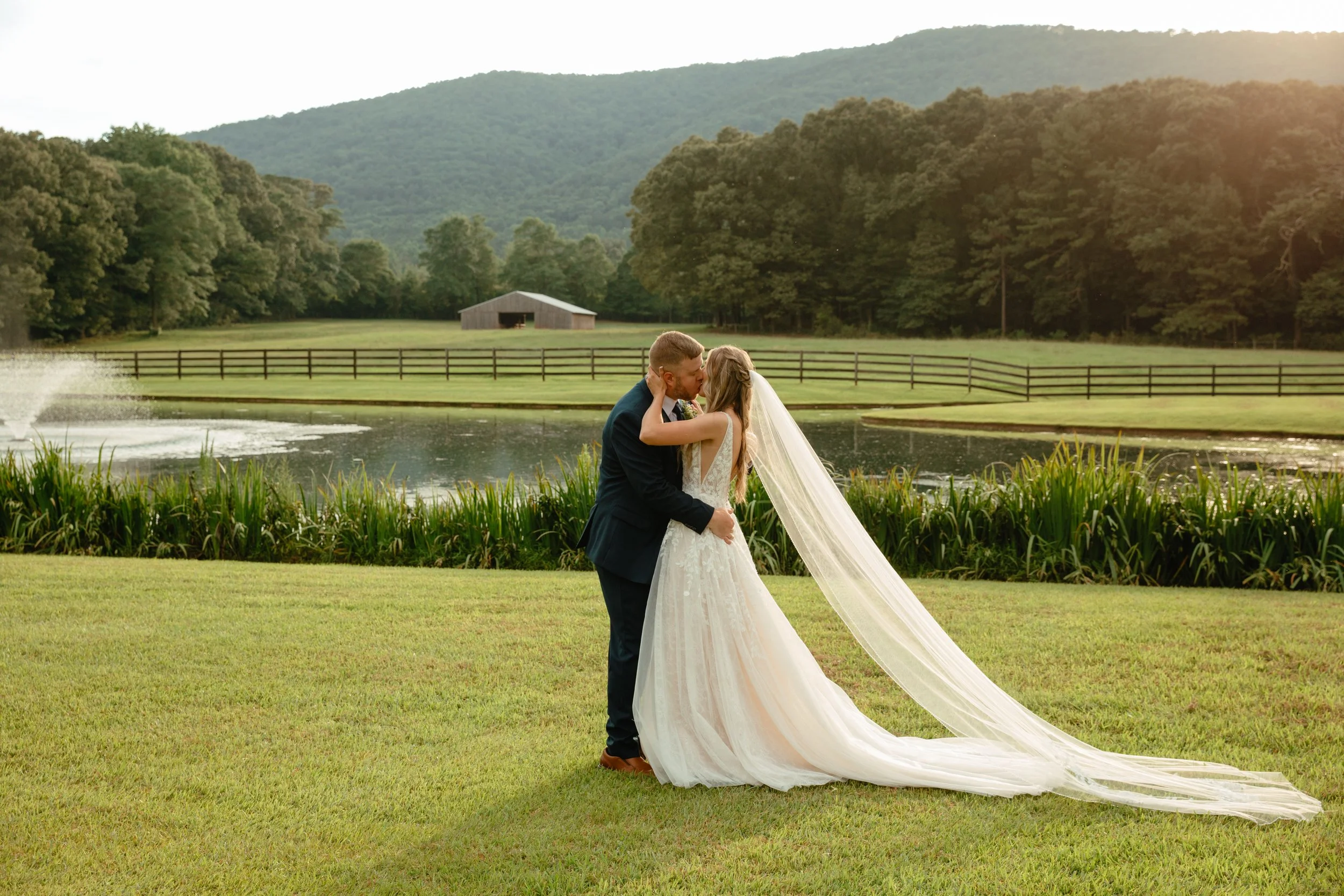 A bride and groom kissing outdoors near a pond, with a wooden fence, trees, and mountains in the background, during what appears to be a wedding photoshoot.
