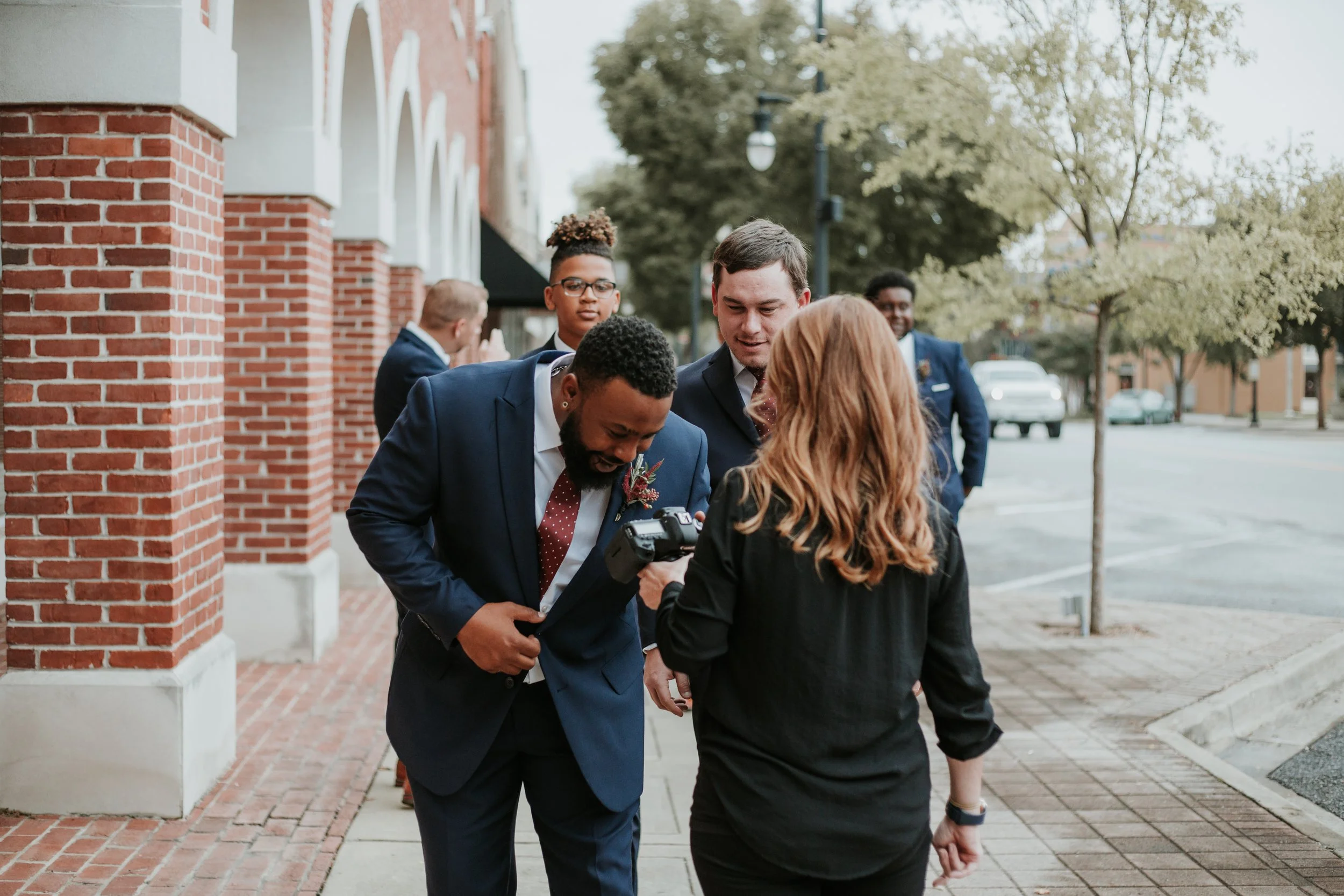 Group of people dressed in formal attire, possibly at a wedding or event, walking along a brick sidewalk with trees and buildings in the background.