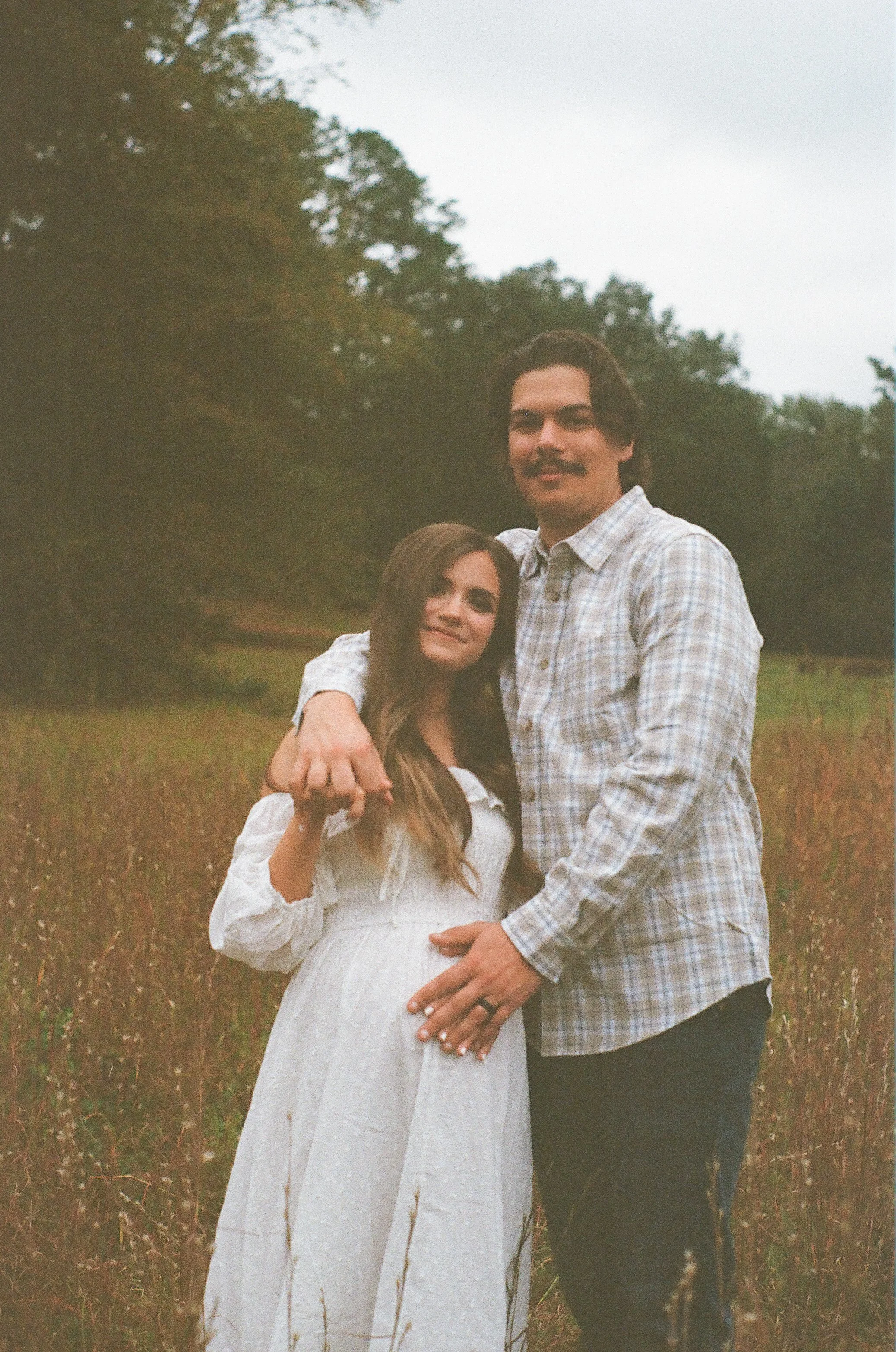 A couple standing in a field, with the man embracing the woman and both smiling at the camera, trees in the background, overcast sky.