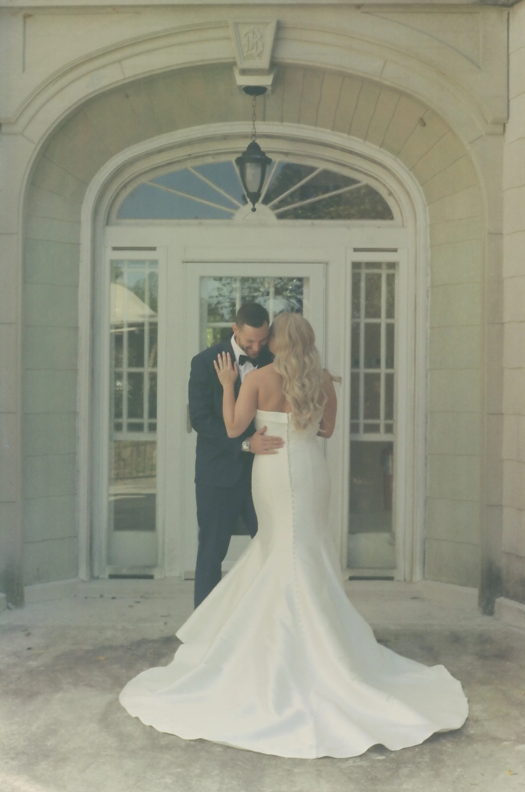 A bride in a white wedding gown and a groom in a black tuxedo share a tender moment in front of a glass door entrance with decorative trim. She has long, blonde, wavy hair, and he has short dark hair.