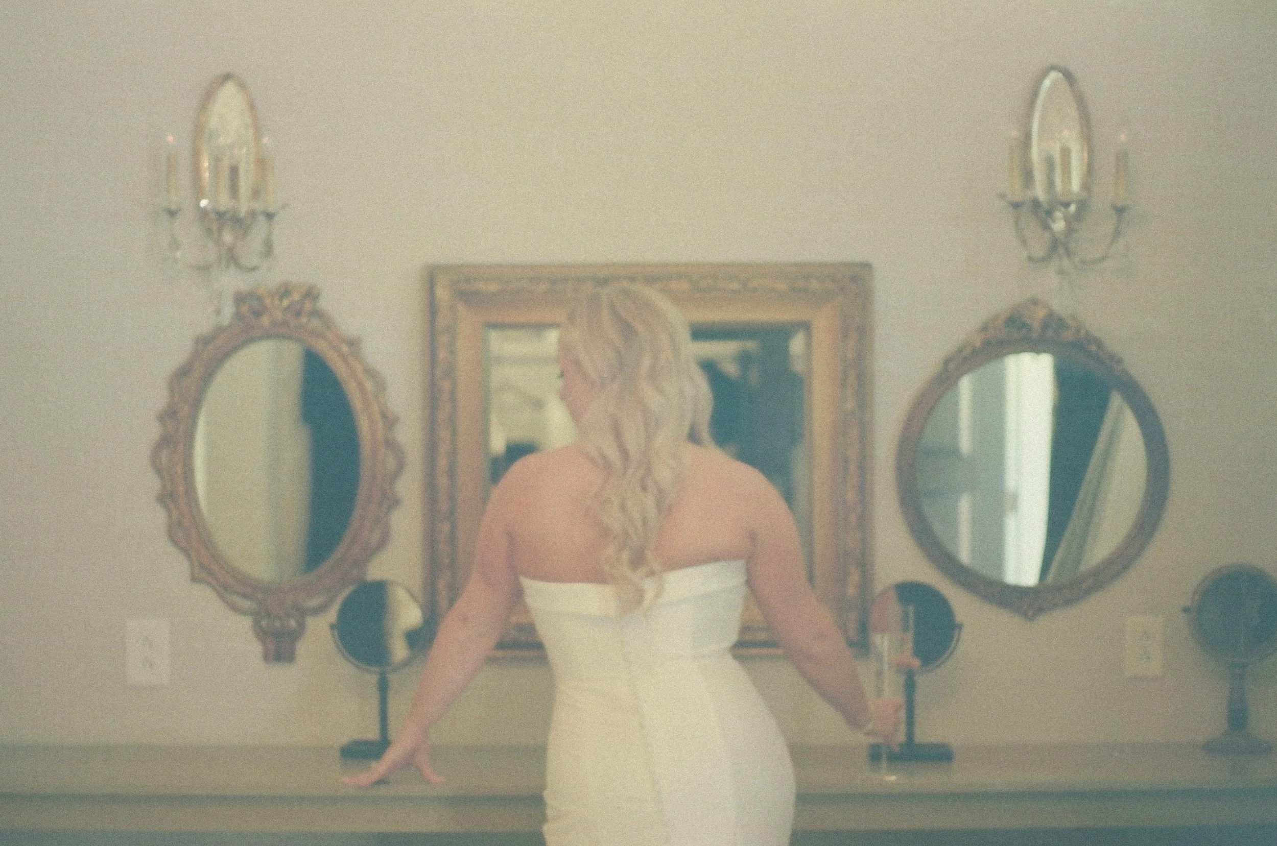 A woman in a white strapless dress standing in front of a mirror with two smaller oval mirrors on either side, a larger square mirror in the center, and wall sconces.
