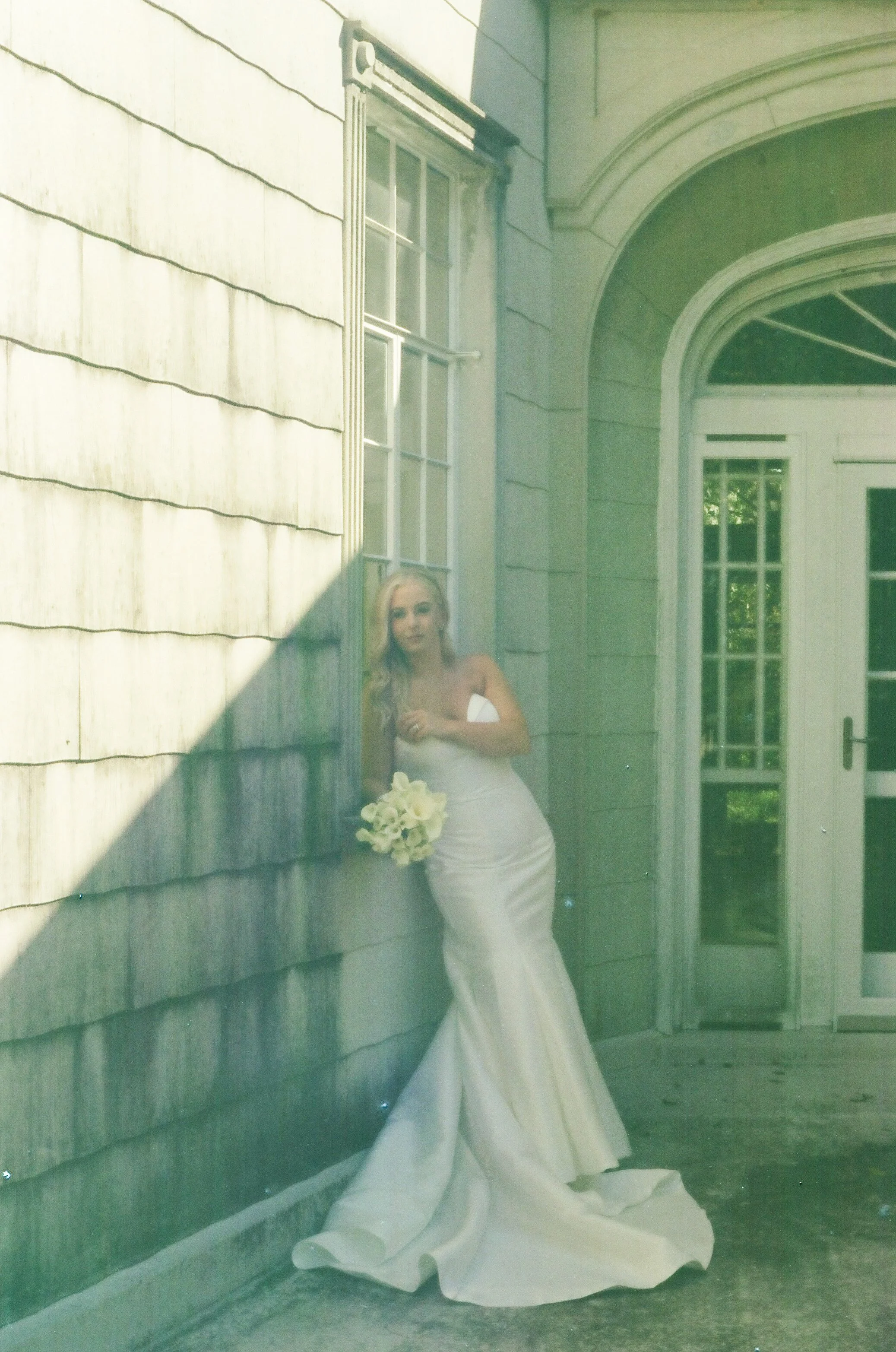 A bride in a white wedding dress holding a bouquet leaning against a wooden wall outside a building with large windows and an arched door.