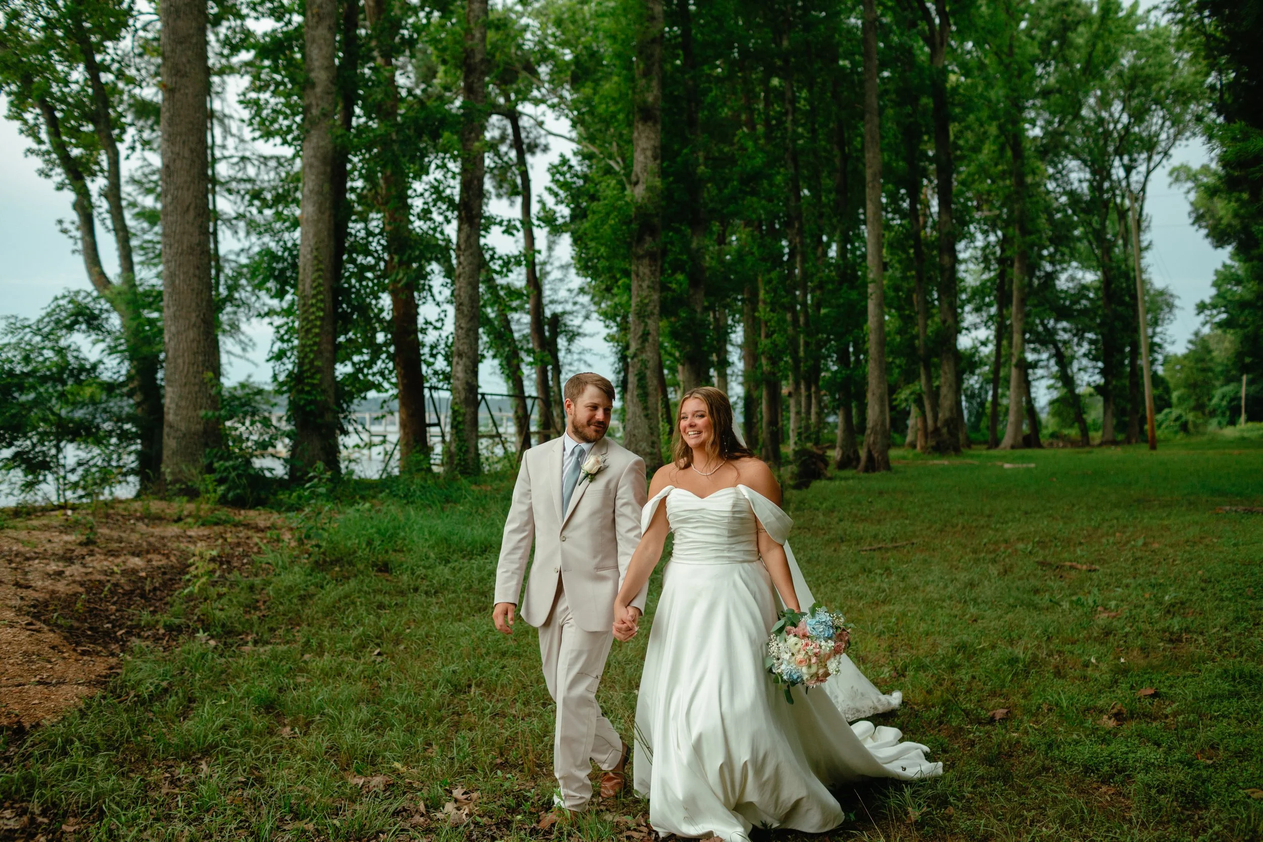 A bride and groom walking hand in hand outdoors in a forested area during daytime, with the bride holding a bouquet of flowers.