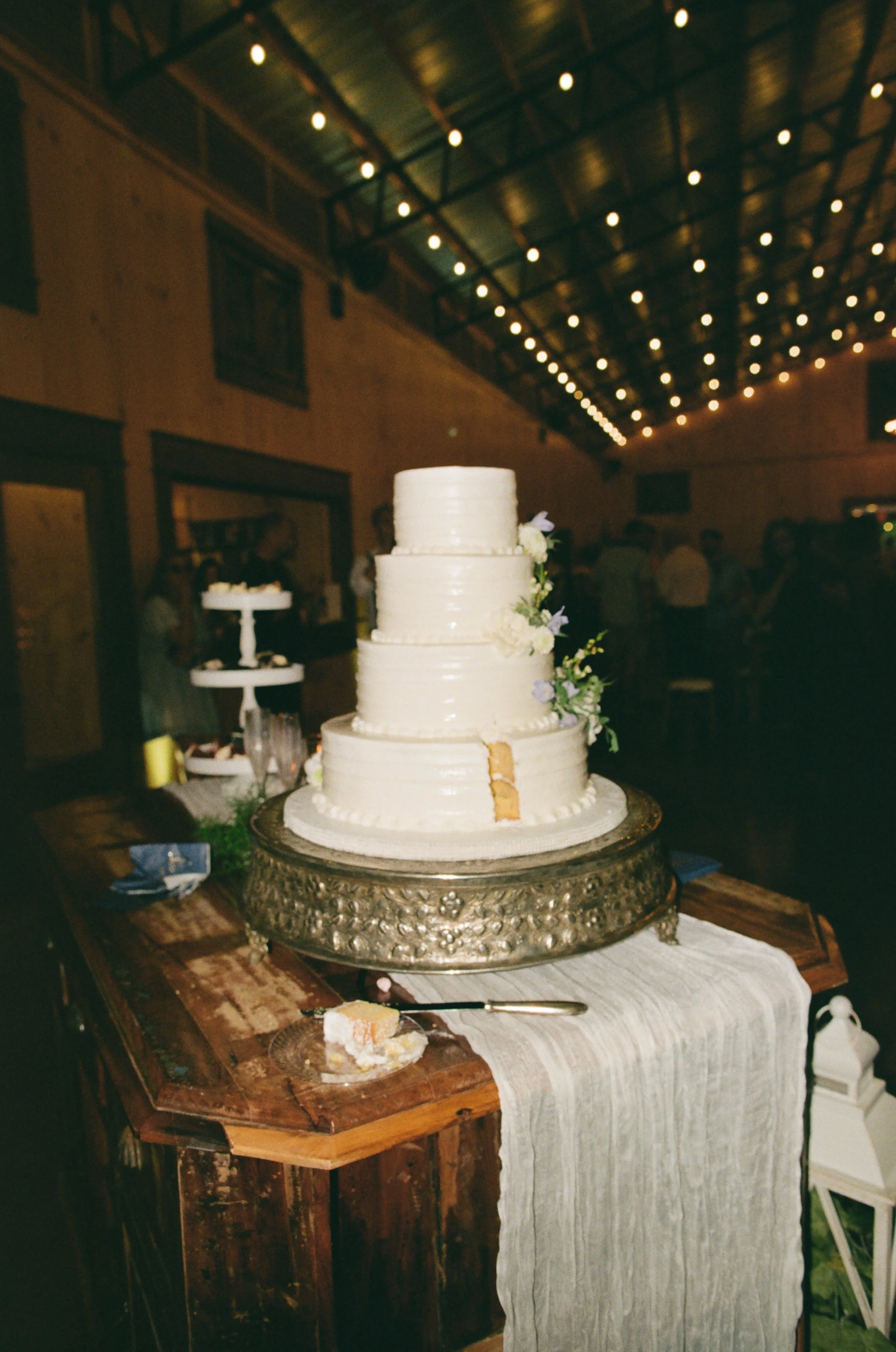 A four-tier white wedding cake with a small piece cut out, decorated with flowers, on a decorative silver cake stand at a wedding reception on 35mm film