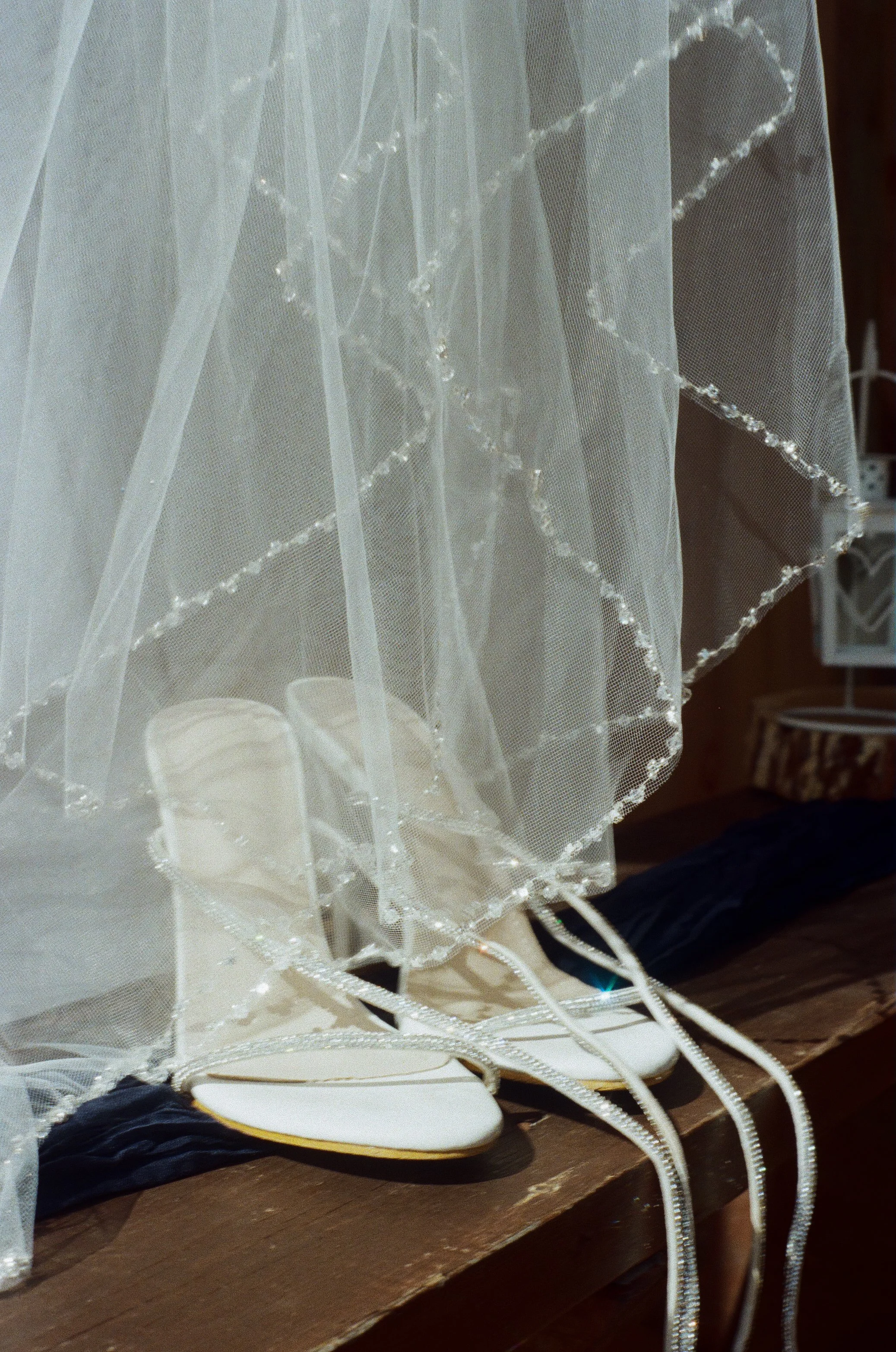 White wedding shoes under a sheer veil with beaded trim on a wooden surface.