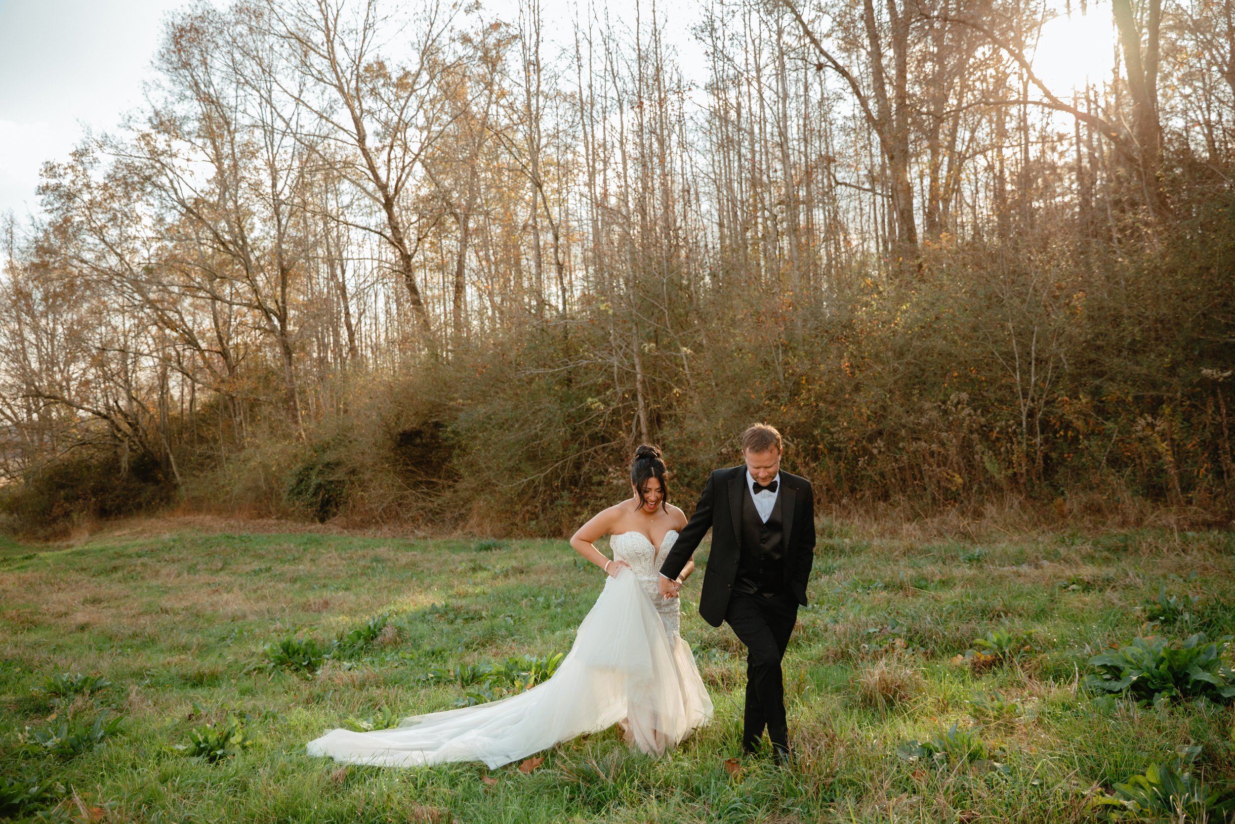 Bride and groom holding hands and walking in a grassy field with trees in the background and sunlight shining through.  Anniston, Birmingham, Heflin, Calhoun Co, Alabama wedding photographer