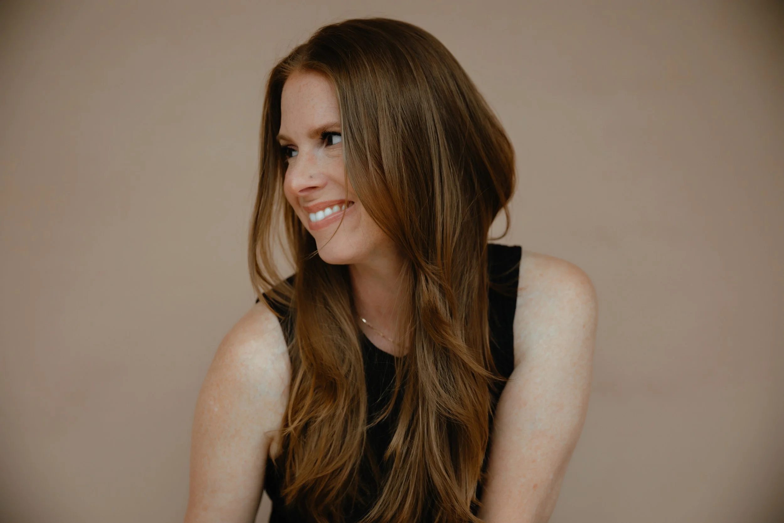 A smiling woman with long, wavy auburn hair and freckles, wearing a sleeveless black top, looking to her left against a plain beige background.