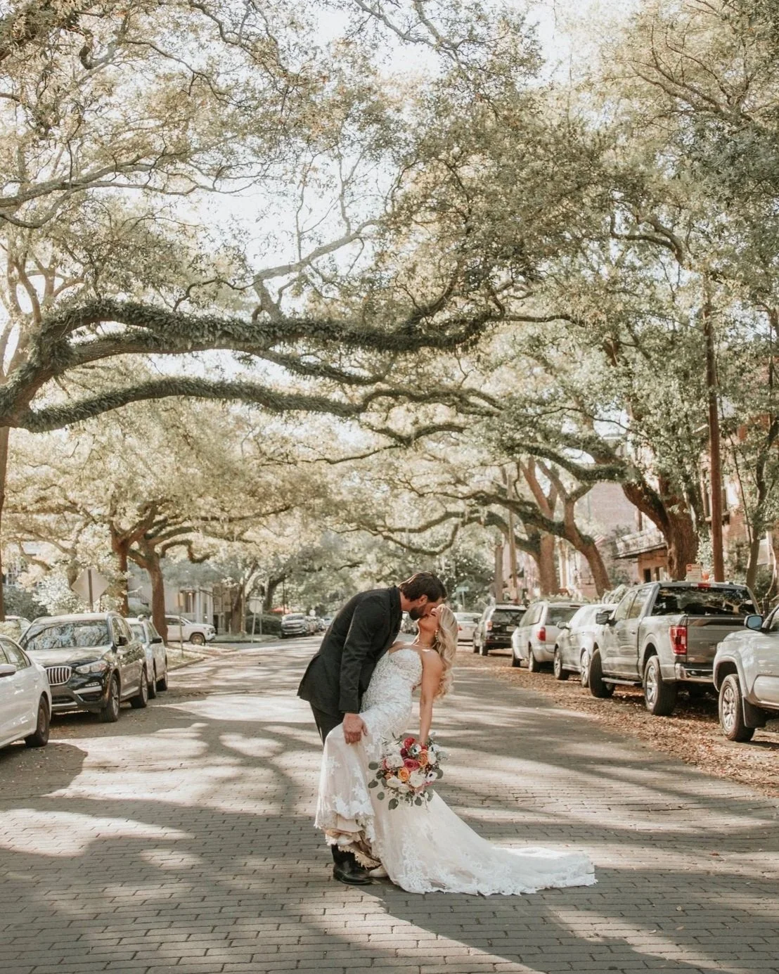 A couple in wedding attire sharing a kiss under large tree branches on a city street, with parked cars along the sides.