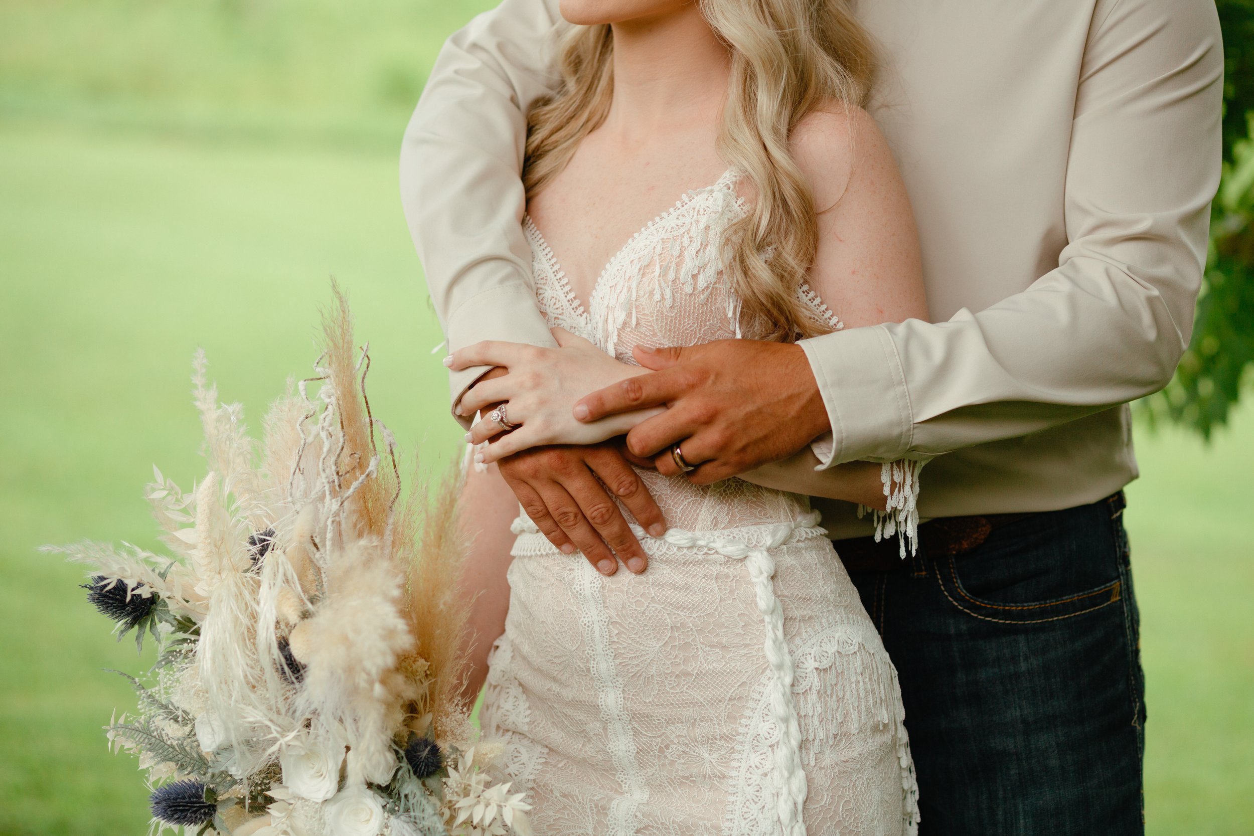 Close-up of a bride and groom during a wedding, with the bride holding a floral bouquet and the groom embracing her from behind in an outdoor setting.