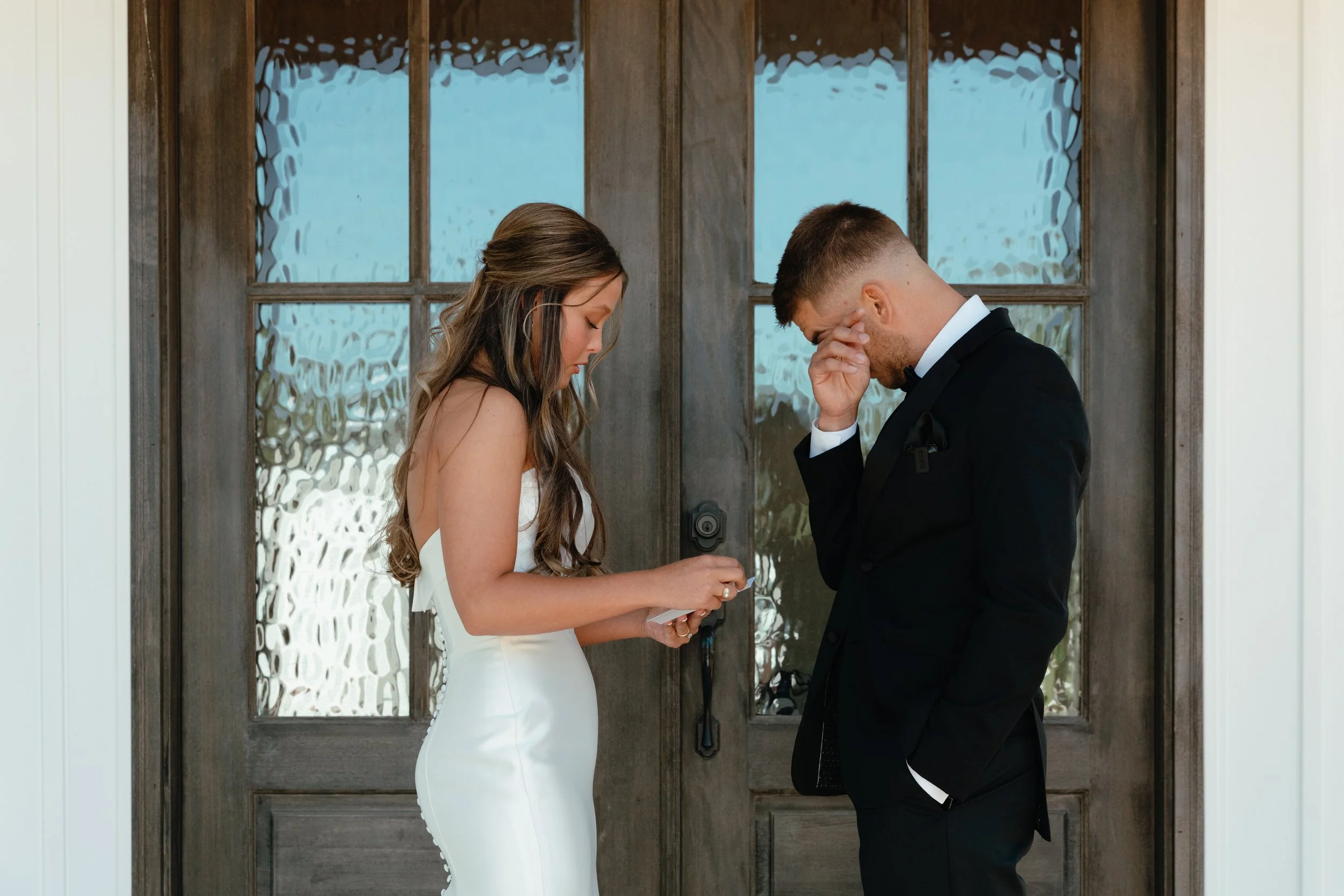 A man and woman stand in front of a glass-paneled wooden door, facing each other with heads bowed and hands raised, appearing emotional during a wedding or ceremony.