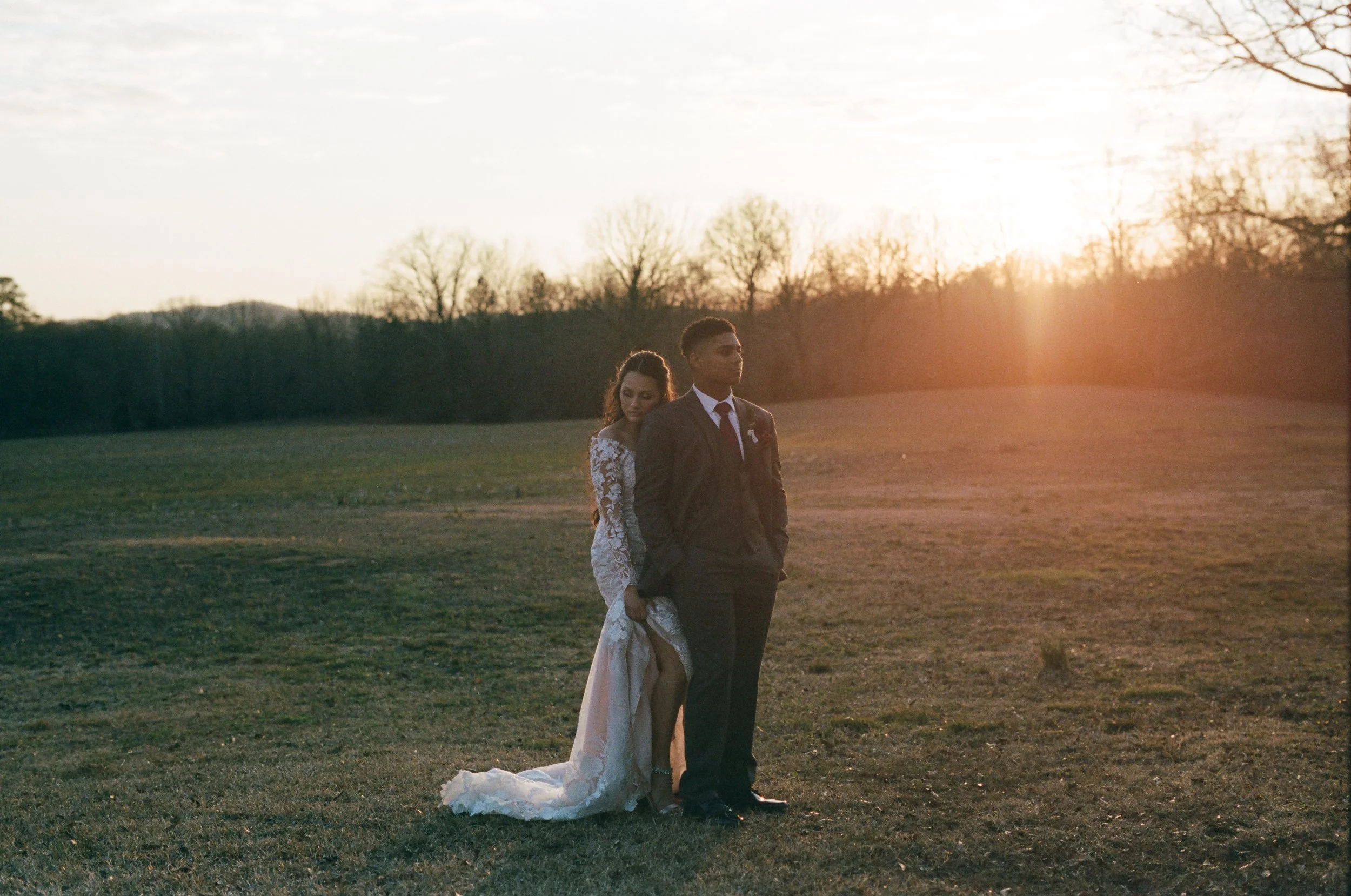 A couple stands outdoors on a grassy field during sunset. The woman is wearing a lace wedding dress and the man is in a dark suit. They appear contemplative, with the woman resting her head on the man's shoulder.