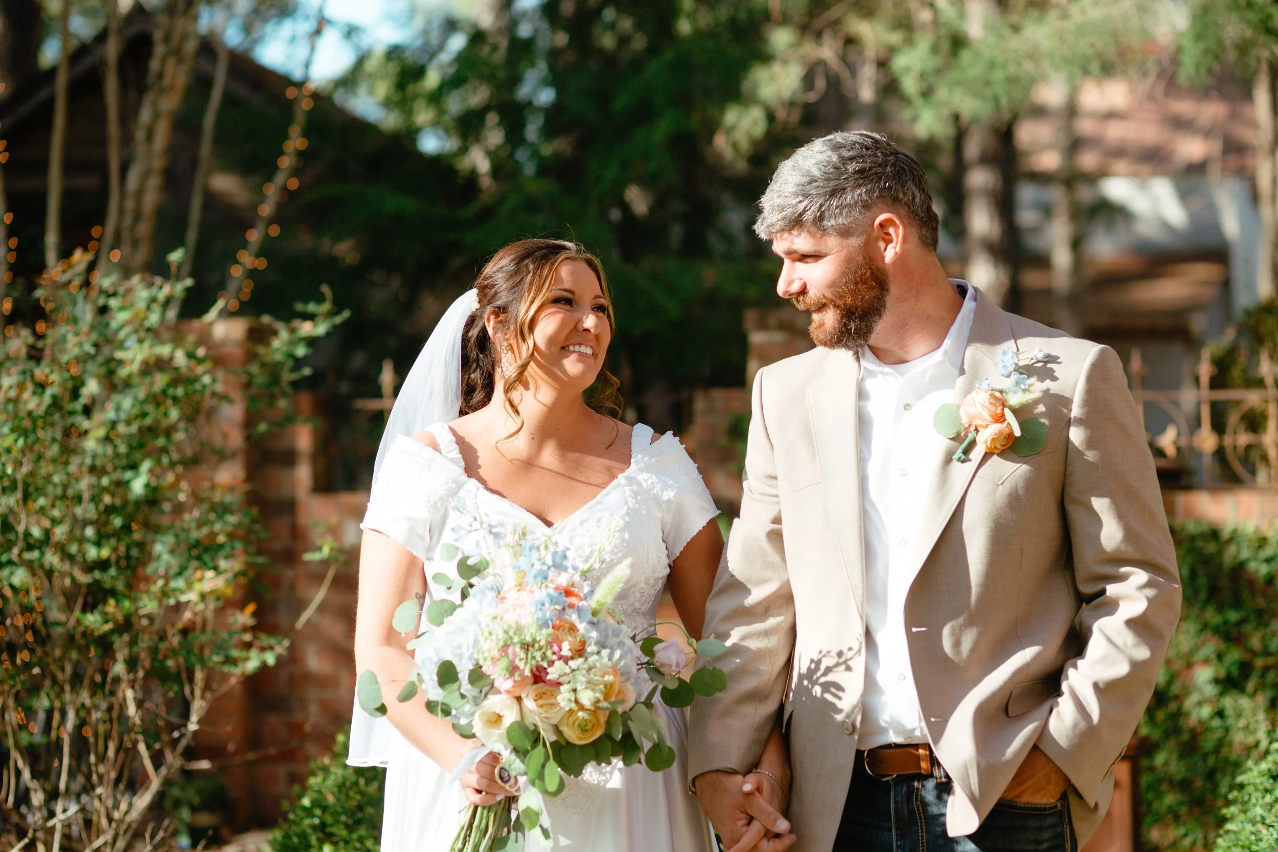 A bride and groom holding hands during an outdoor wedding ceremony, smiling at each other, with trees and string lights in the background.