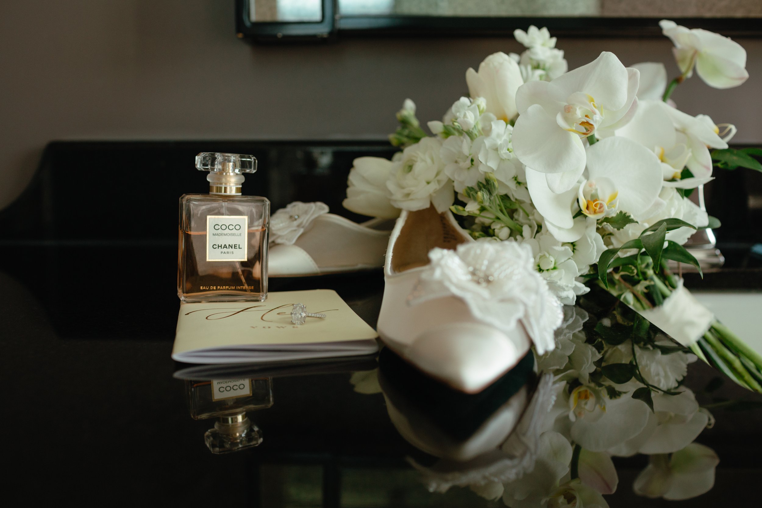 A decorative scene with a perfume bottle labeled 'COCO MADEMOISELLE' from Chanel, an engagement ring resting on a cream-colored card, and a bouquet of white flowers on a reflective black surface.